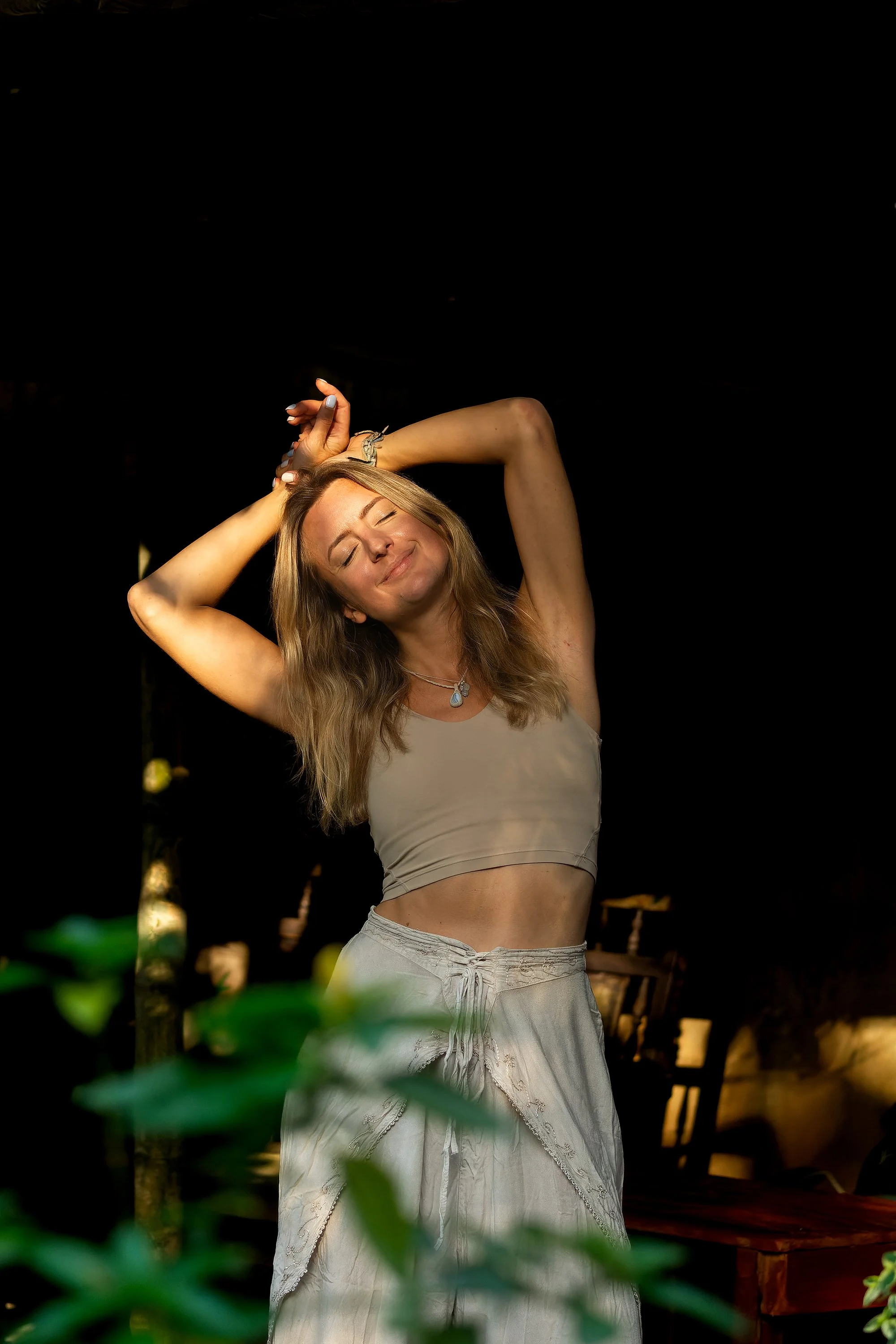 A woman with long hair, wearing a beige crop top and beige pants, is smiling with her eyes closed, standing outdoors at night, surrounded by greenery and furniture.