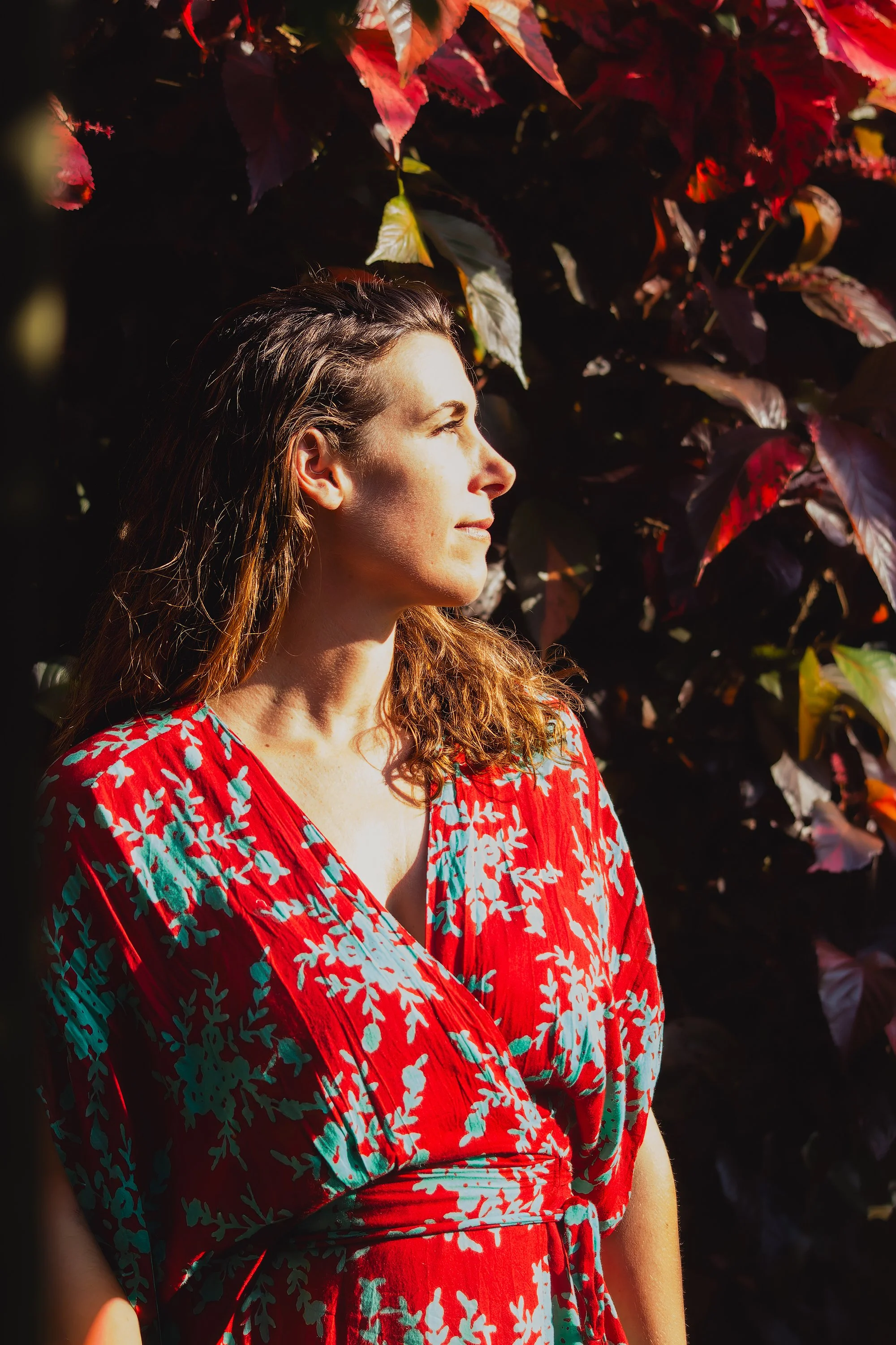 A woman with long hair wearing a red dress with a white floral pattern, standing outdoors in front of dark green and burgundy leaves.