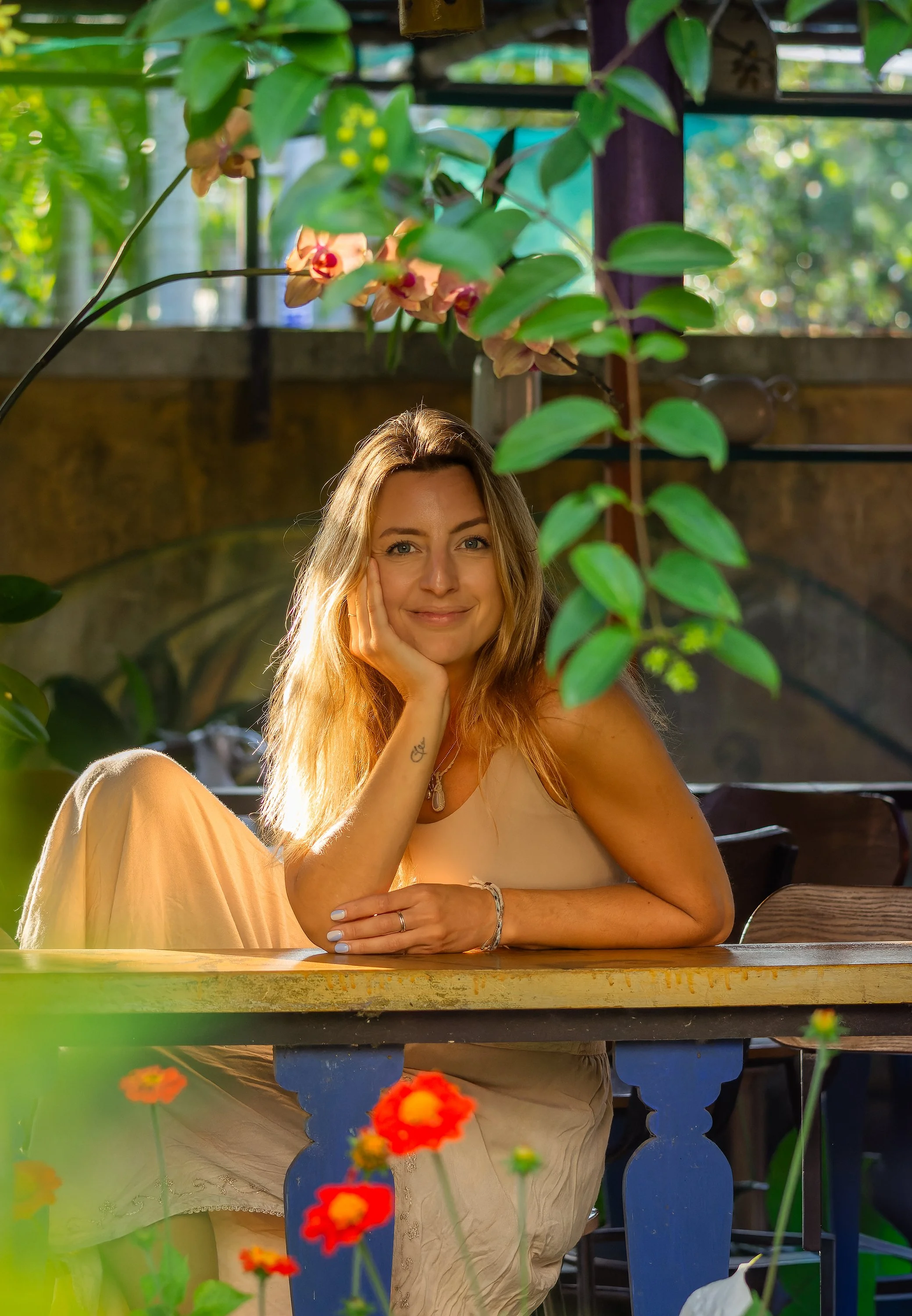 A woman with long, wavy hair sitting at a wooden table in a lush, green environment. She is resting her head on her hand, smiling softly, surrounded by vibrant flowers and foliage.