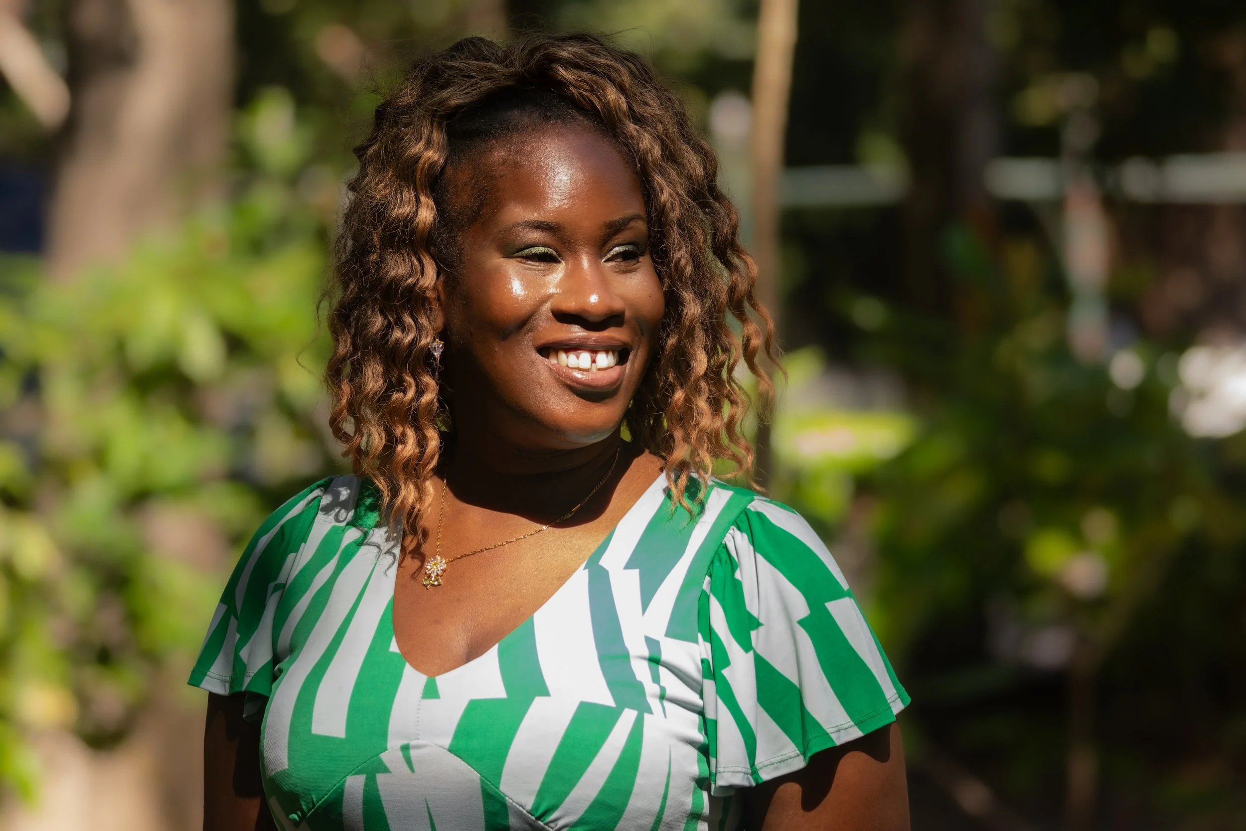 A woman with curly brown hair, smiling outdoors in a green and white patterned top with a black collar, in a natural setting with trees and sunlight.