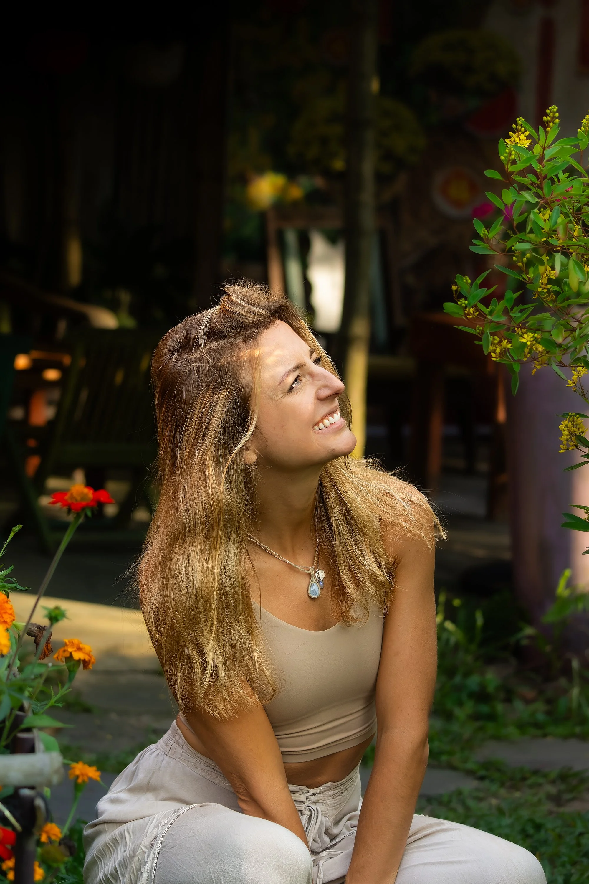 A woman with long blonde hair smiling and looking upwards in a garden with colorful flowers and greenery.