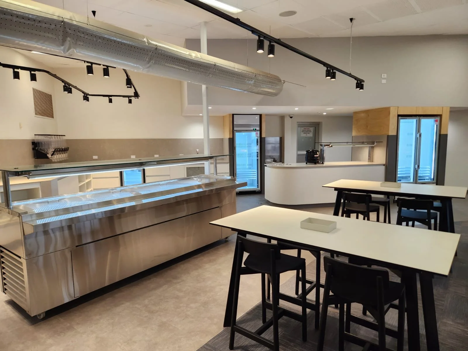 Empty cafeteria or dining area with tables and chairs, a stainless steel serving counter, refrigerator, and kitchen appliances in the background.