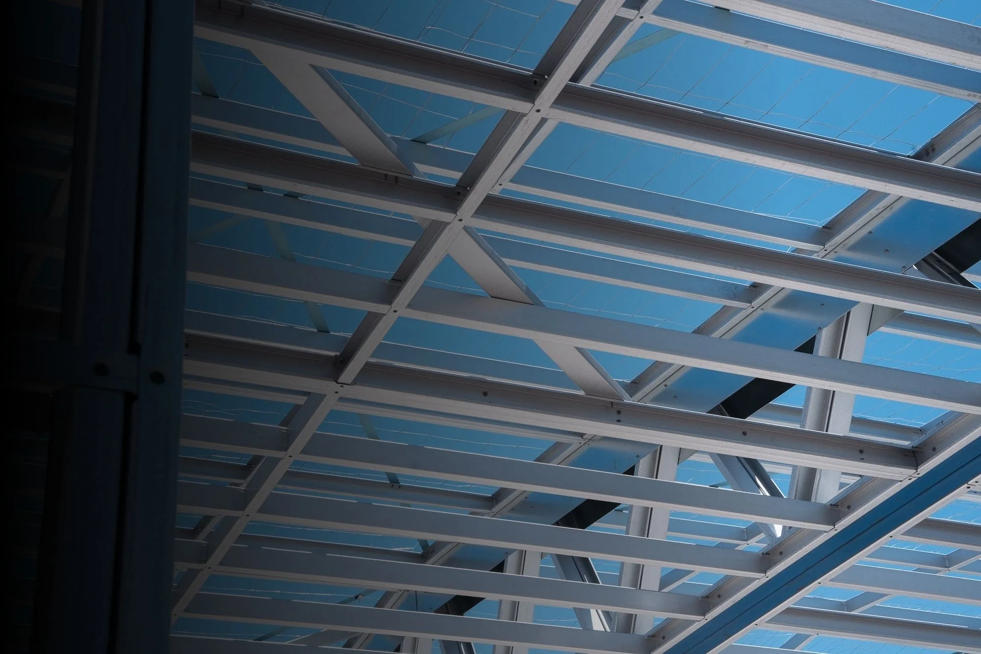 Looking up at a modern glass and metal roof structure with a geometric pattern against a clear blue sky.