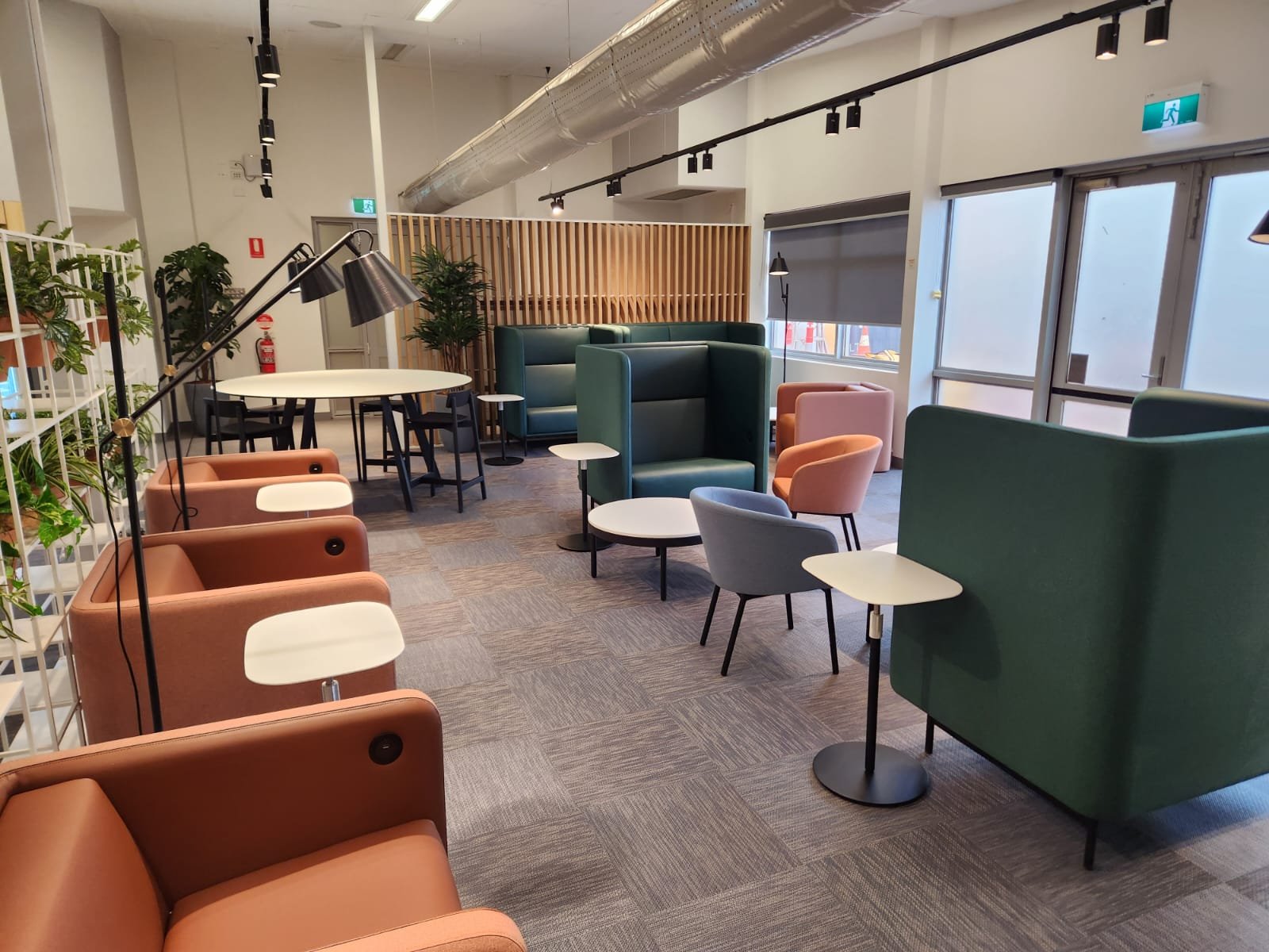 An indoor lounge or waiting area with colorful armchairs, small side tables, and a large window with blinds, decorated with plants and modern furniture.
