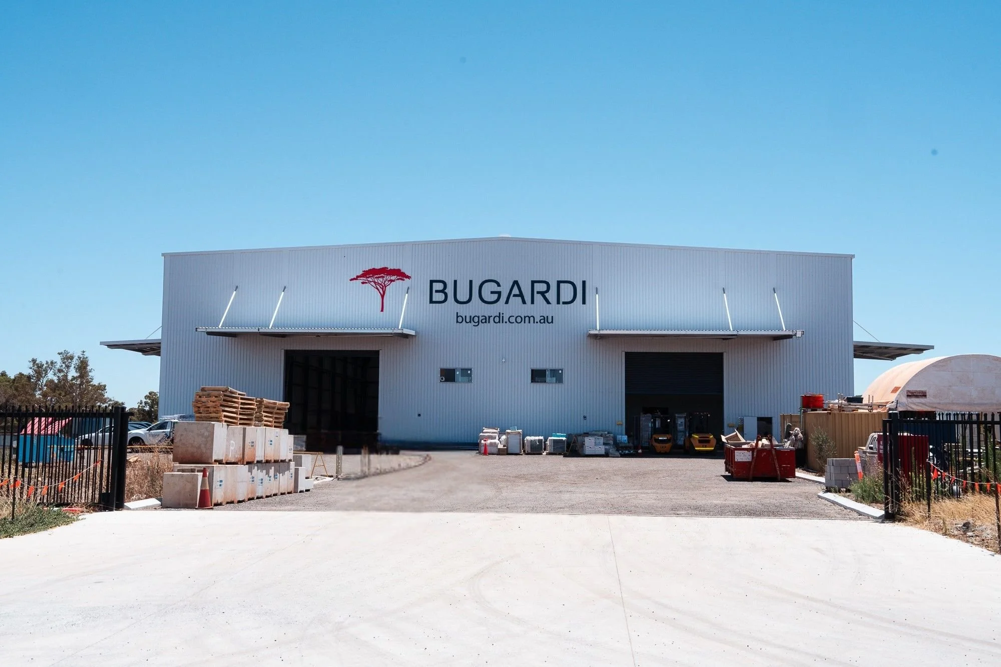 Front view of a large warehouse with a sign that reads 'BUGARDI' and the website 'bugardi.com.au.' The warehouse is situated in an industrial area with various construction materials, pallets, and vehicles around it. The sky is clear and blue.
