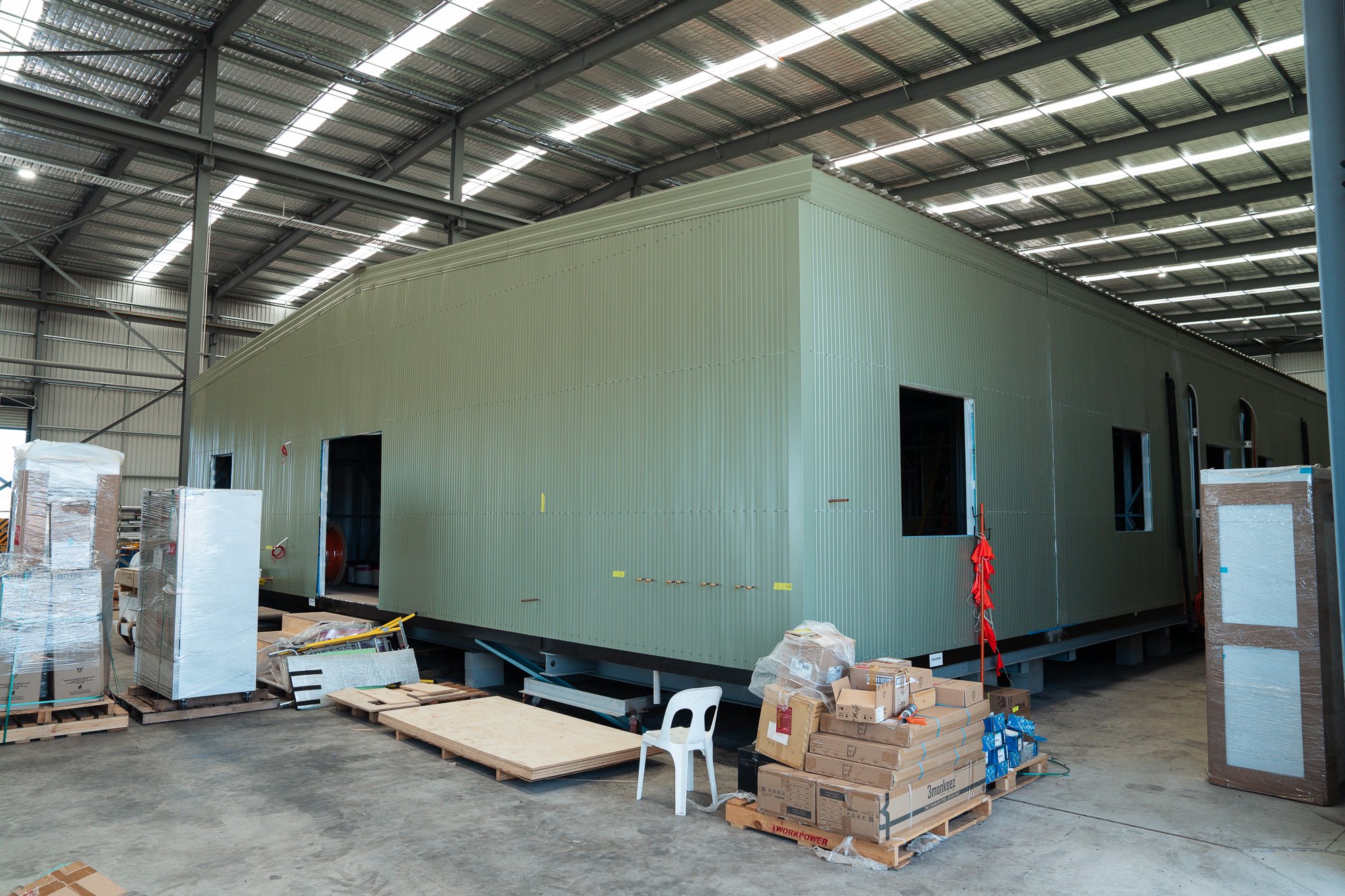 Interior of a warehouse with a partially assembled green prefabricated building, surrounded by boxes, pallets, and construction materials.