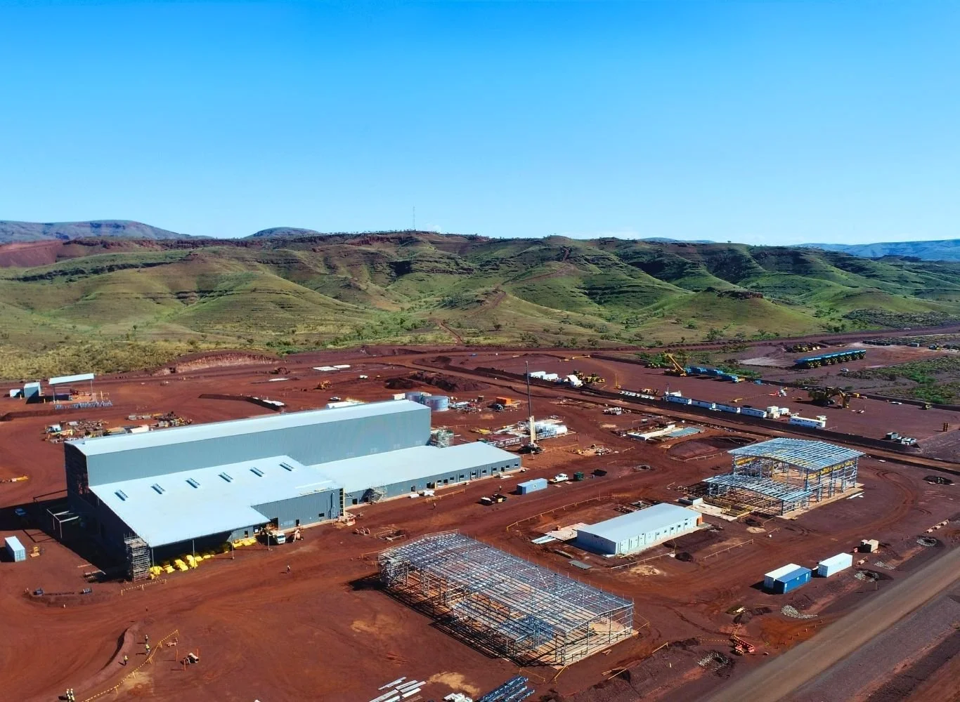 Construction site with large white warehouse, metal frameworks, and dirt ground, set against green hills and blue sky.