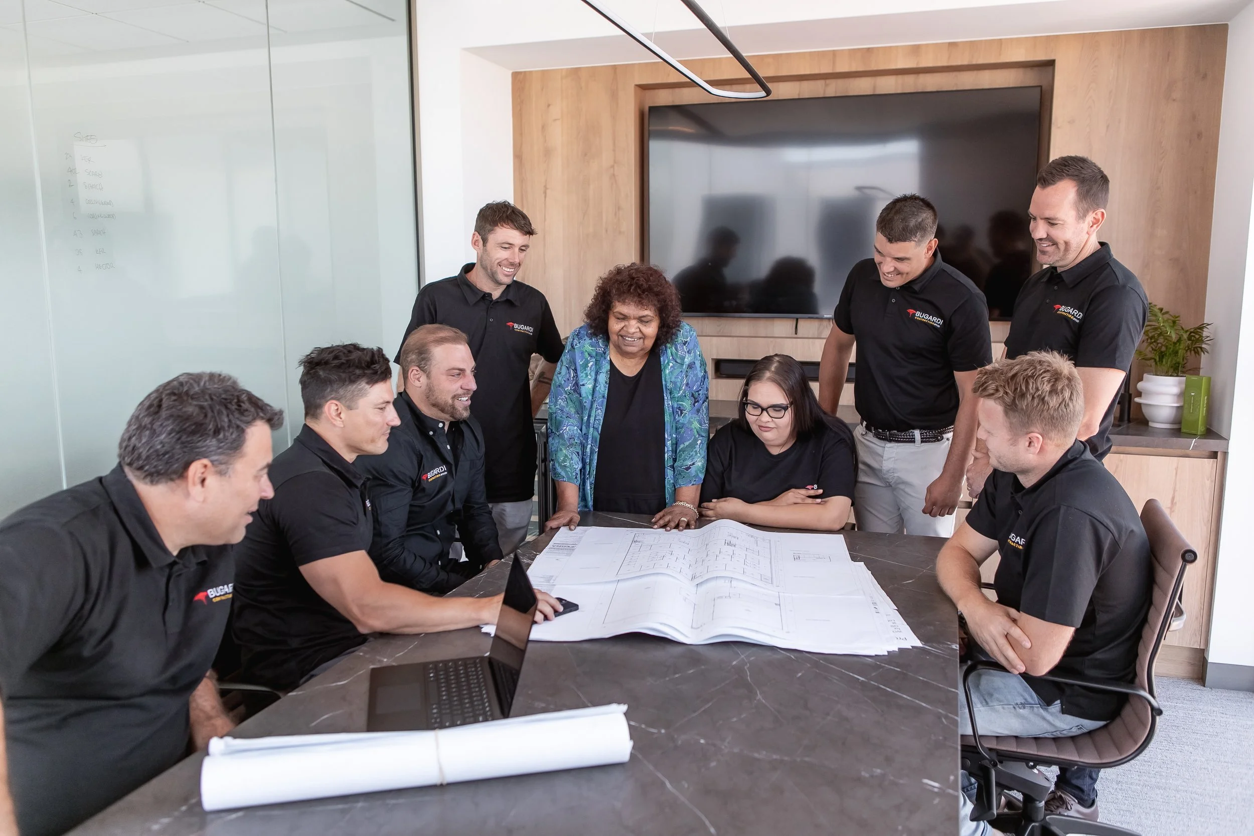 A group of diverse professionals gathered around a table in a modern conference room, engaging in a discussion. Some are seated, and a woman and three men are standing, all smiling. The table has blueprints, a laptop, and notebooks. The background fe
