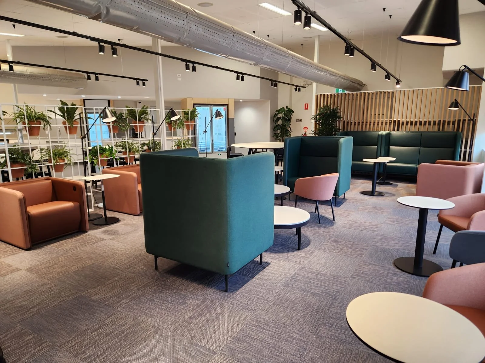 Empty modern lounge with colorful armchairs, small round tables, green plants, and a wooden slatted divider.