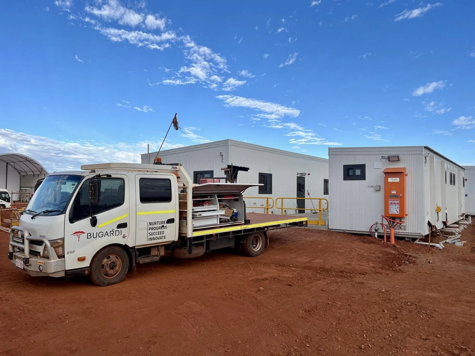 A small white utility truck with the logo 'BUGARDI' and several open compartments, parked on red dirt in front of prefabricated modular buildings under a partly cloudy sky.