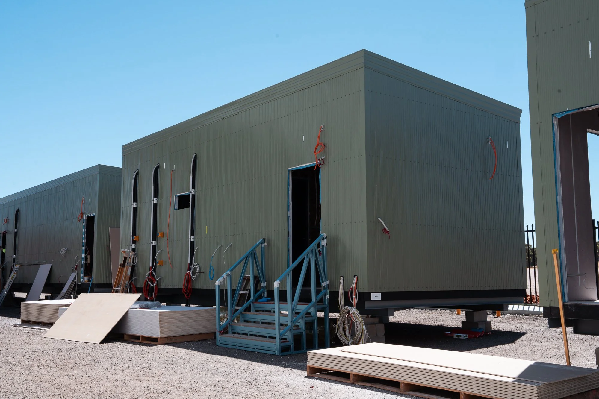 Construction site with several green modular units being assembled, with tools, electrical wires, and building materials around, under a clear blue sky.