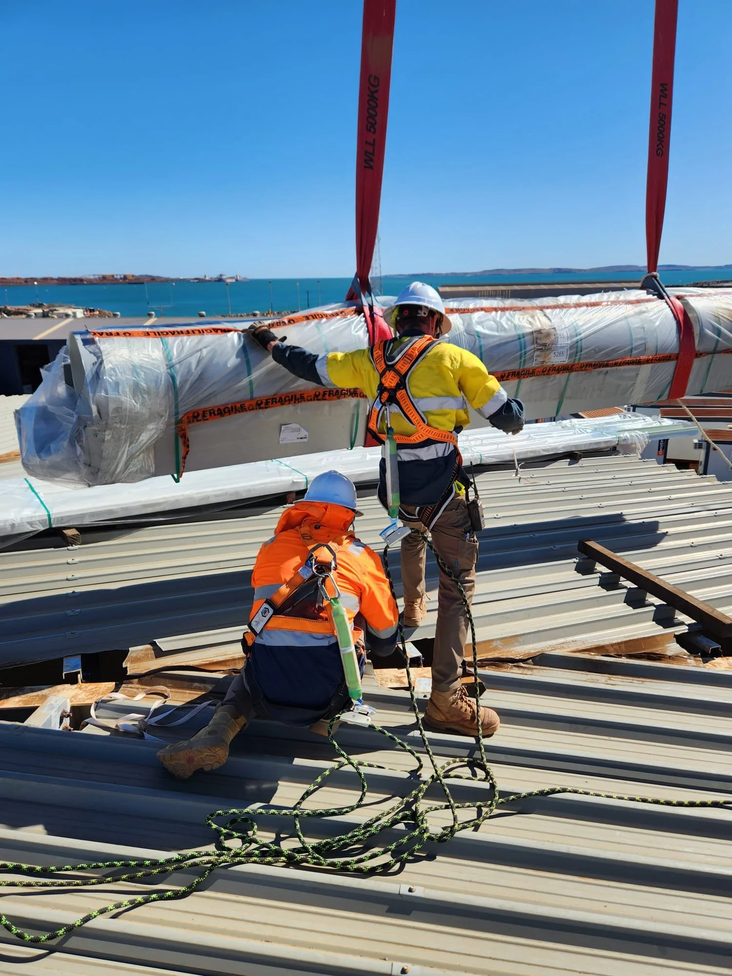 Workers installing or inspecting a wind turbine blade on a metal rooftop near the ocean, wearing safety gear and harnesses.