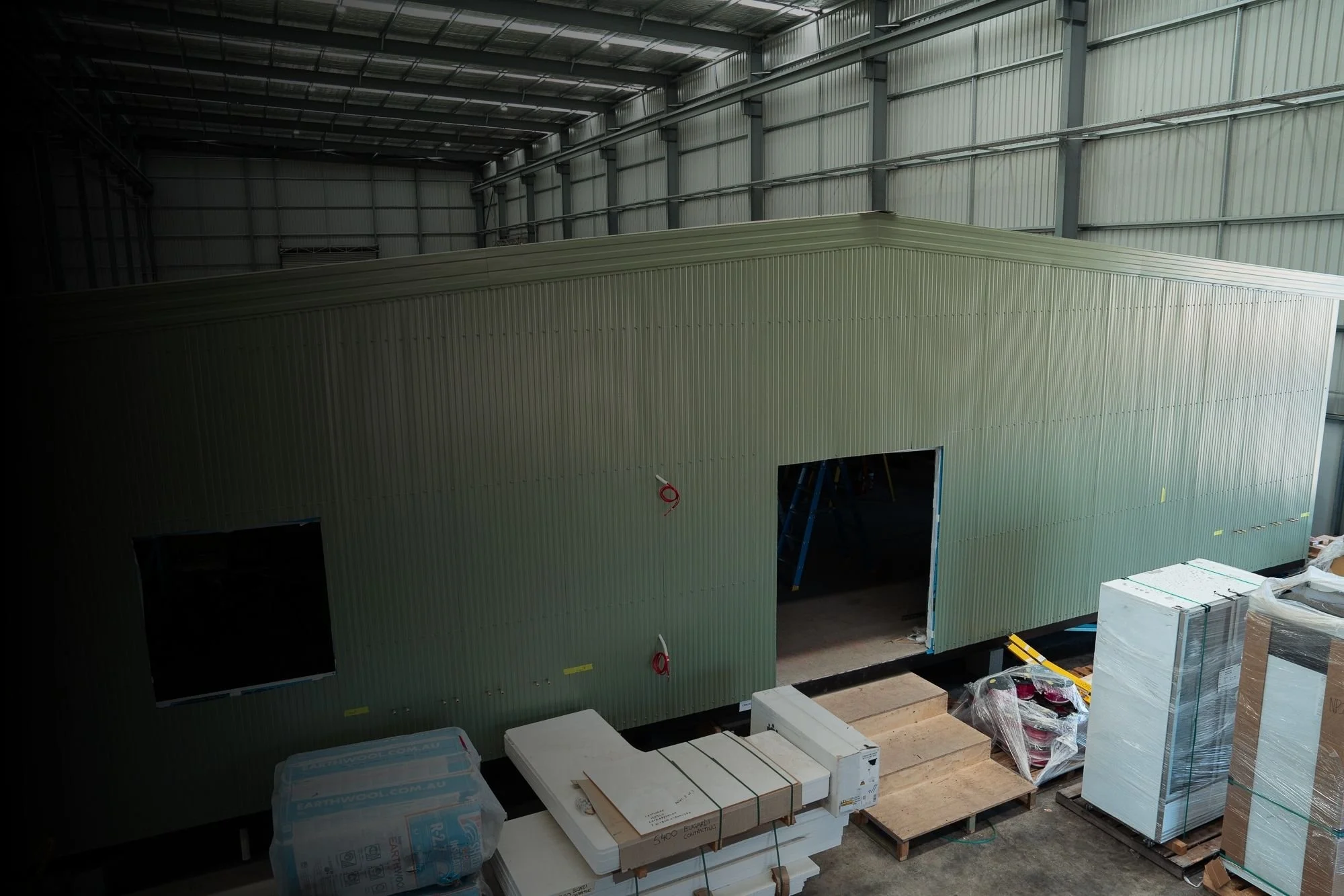 Construction site inside a large warehouse with a partially built green modular structure, packages, and construction materials scattered around.