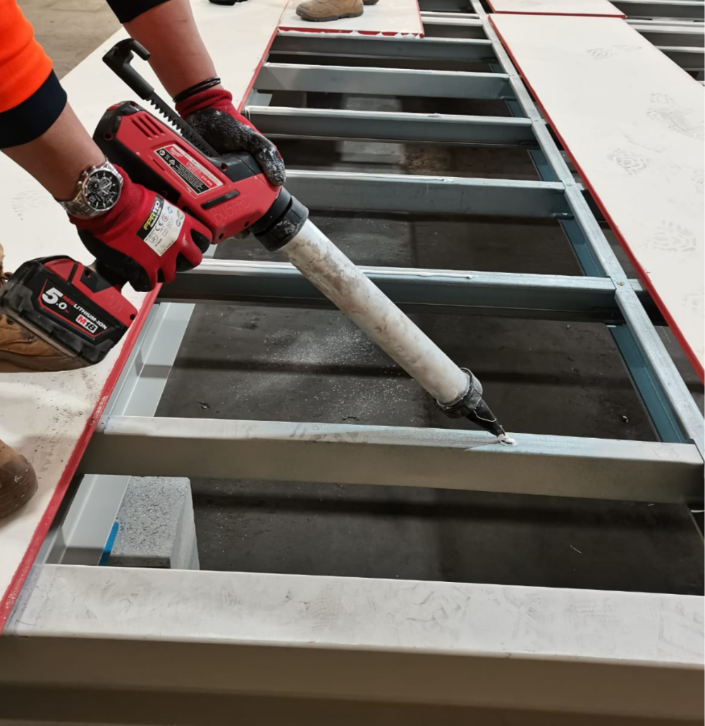 A person using a red and black cordless power tool to weld or spray paint a metal frame on a construction site. The person is wearing red gloves and a wristwatch, with another foot visible in the background.
