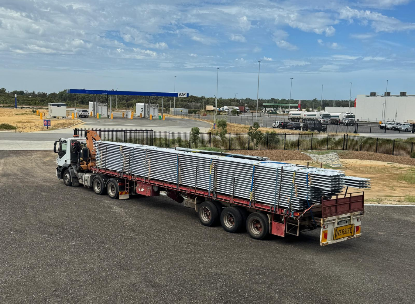 Large flatbed truck carrying metal pipes parked in a construction or industrial area with a gas station in the background.