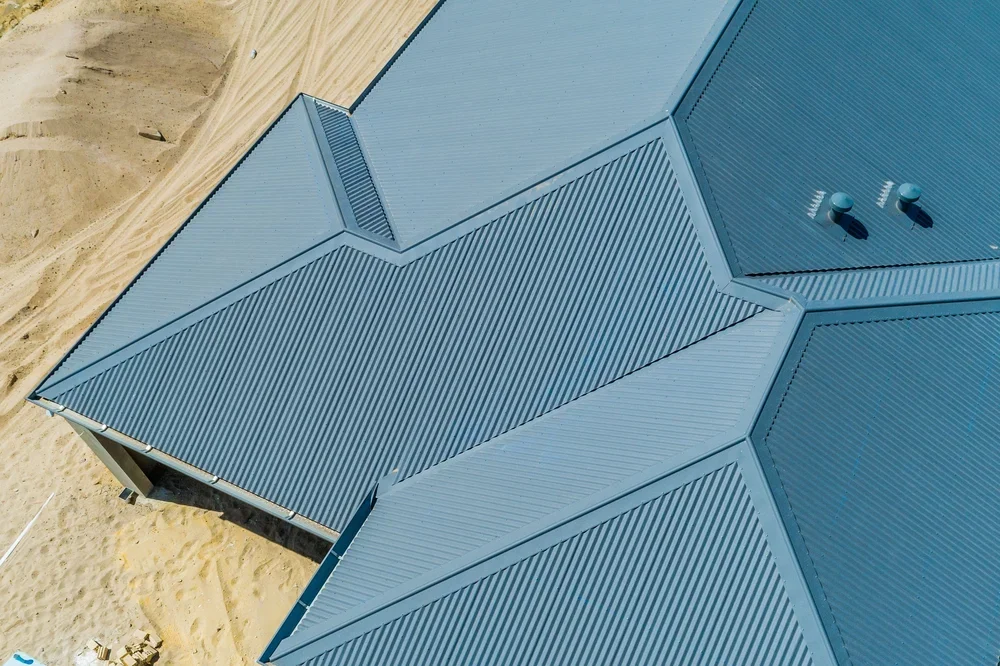 Aerial view of a house with a silver metal roof on sandy ground.