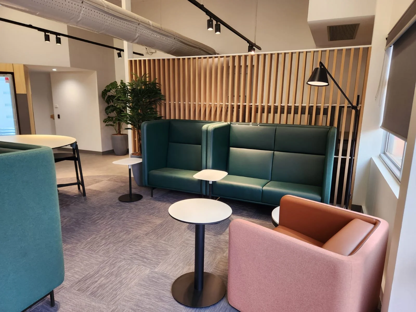 Modern lounge area with green and pink armchairs, small white tables, ample sunlight, and wooden slat divider.