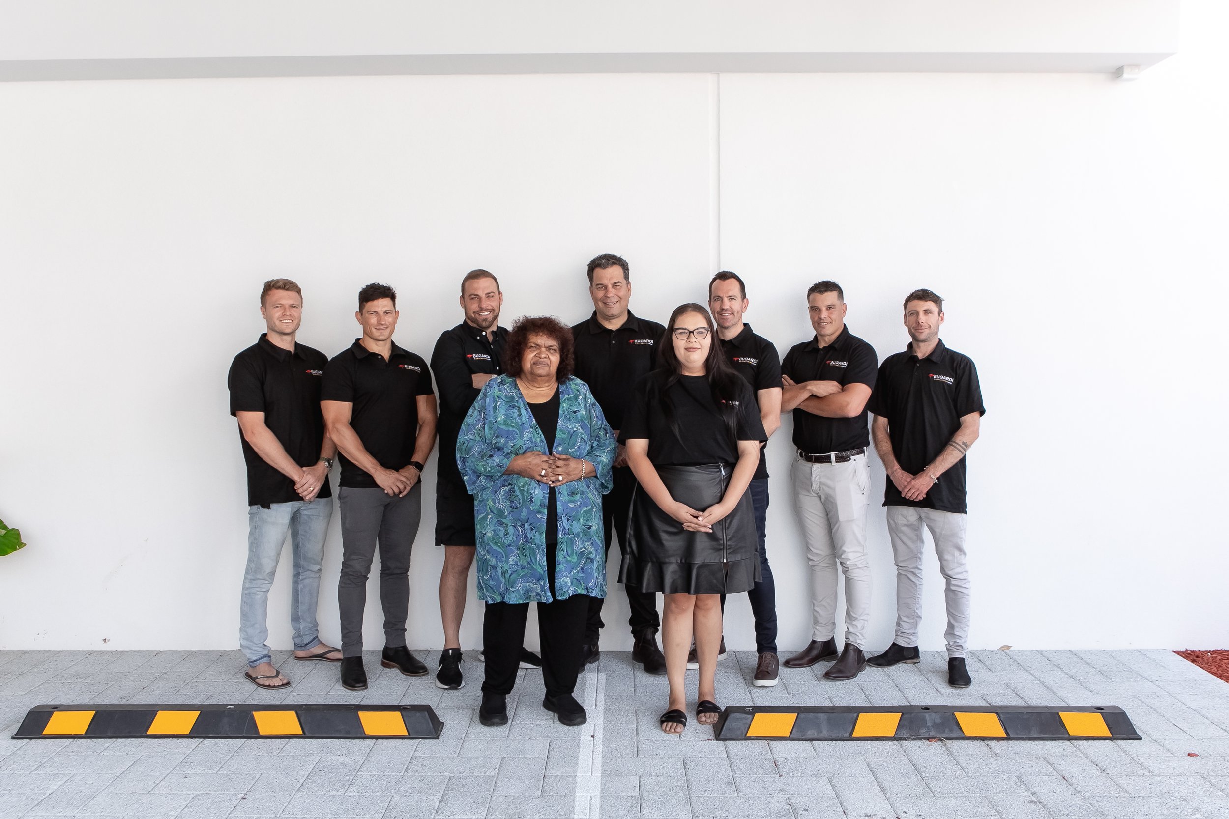 Group of nine people, mostly young men in black shirts and two women, standing in front of a plain white wall with a black and yellow caution strip on the ground.