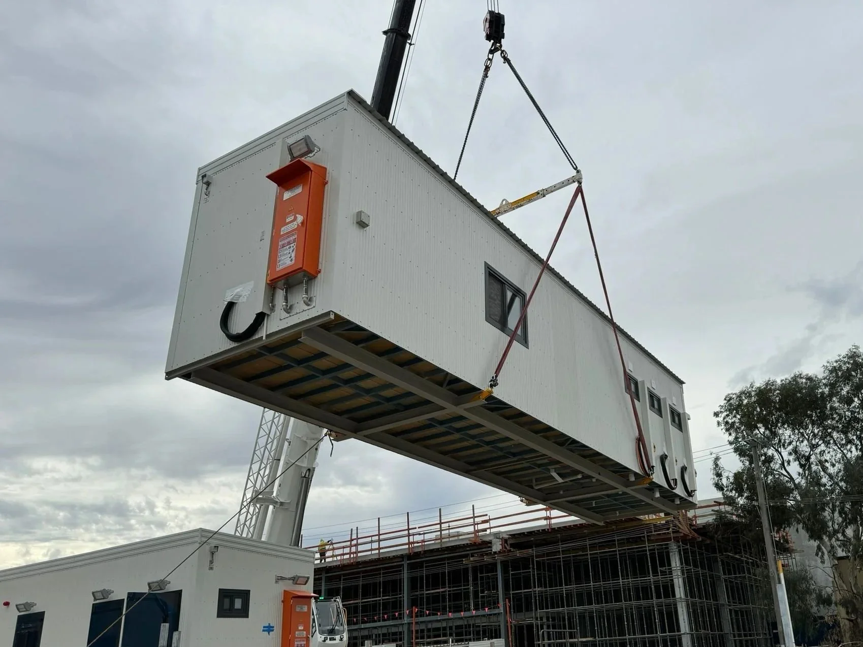 A large modular building section is being lifted by a crane on a construction site. The section has a window and is white with an orange box attached to its side. The sky is cloudy, and there is scaffolding and other construction equipment nearby.