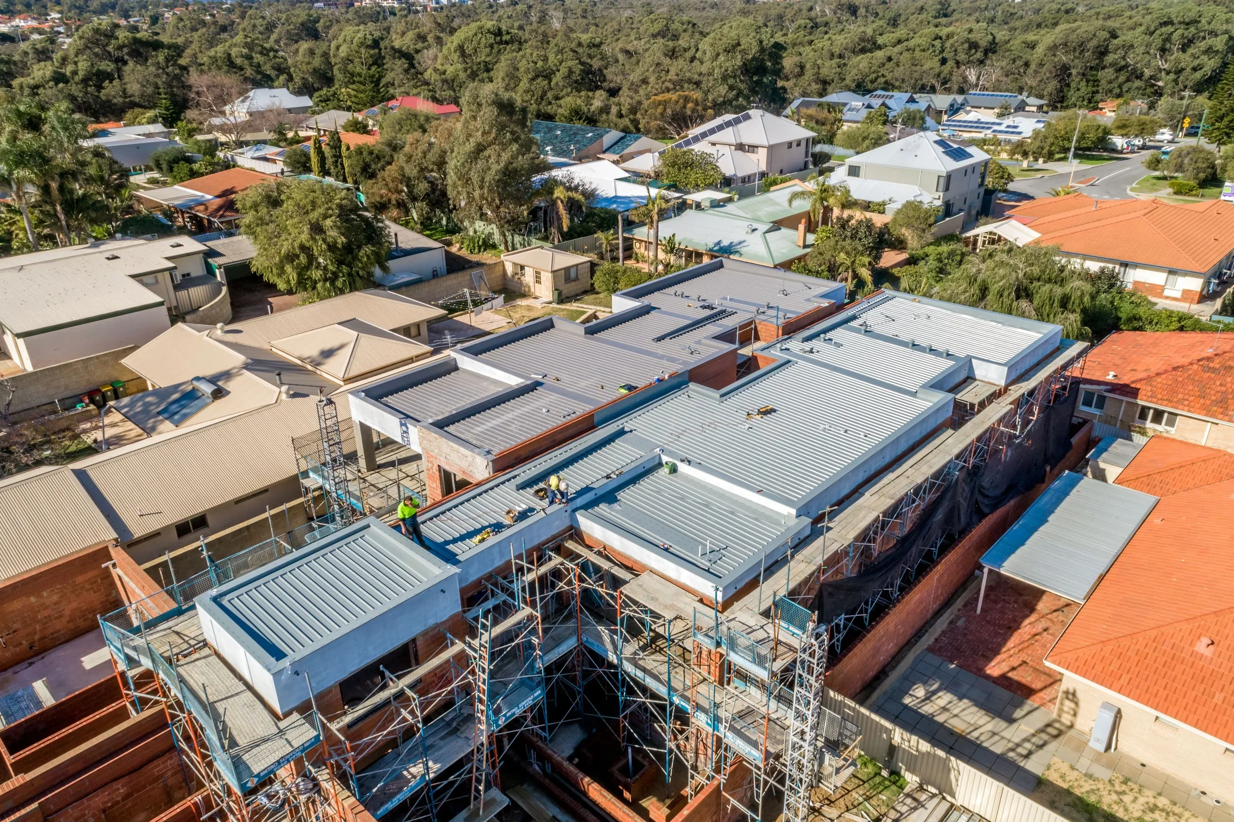 An aerial view of a residential neighborhood with a construction site where workers are installing a metal roof on a building, surrounded by trees and other houses.