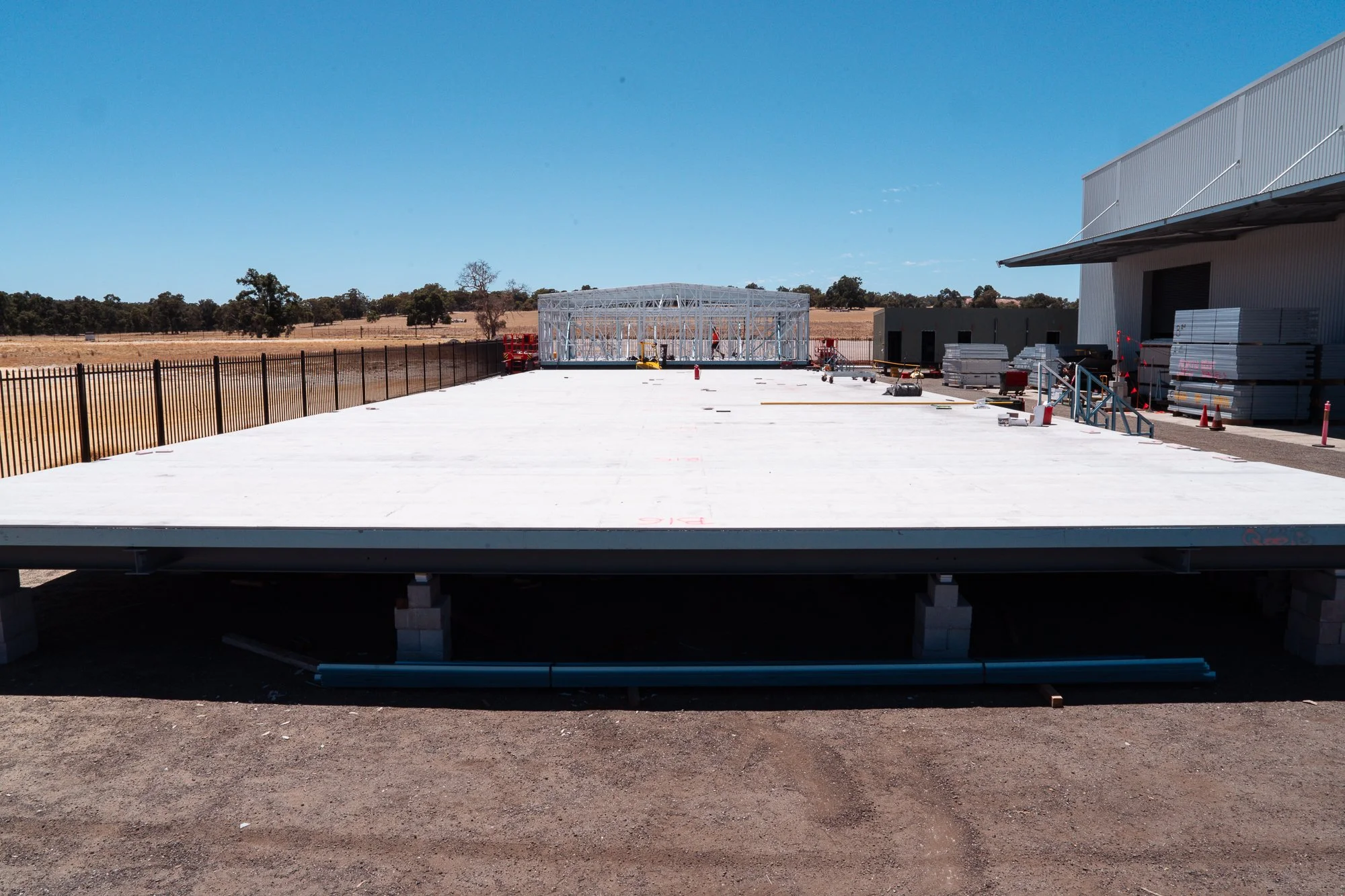 Construction site with a large concrete slab foundation, metal frame structure, and a warehouse building nearby under clear blue sky.