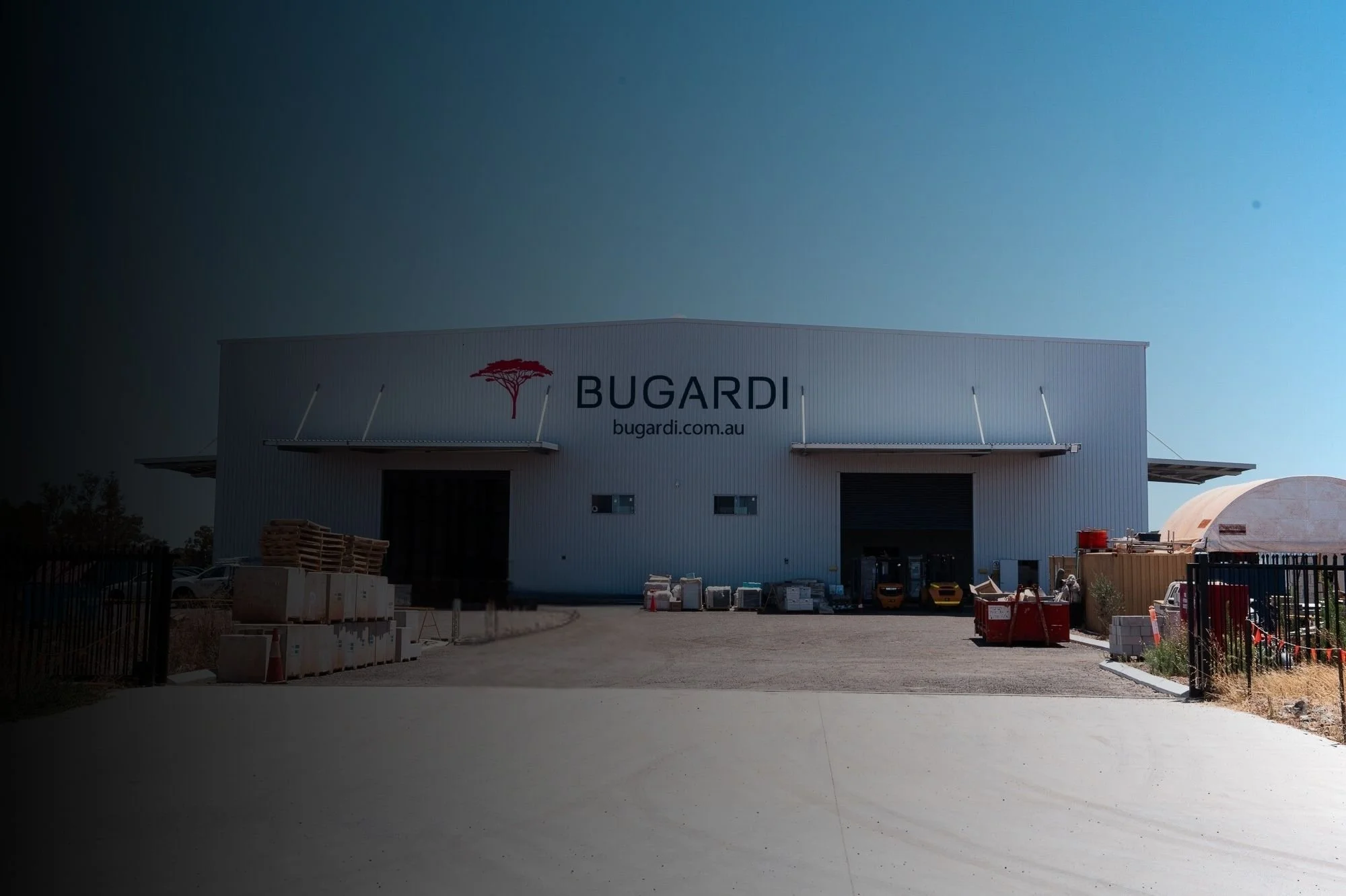 Exterior view of a large warehouse building with the logo and website "BUGARDI" on it, surrounded by construction materials and equipment, under a clear blue sky.