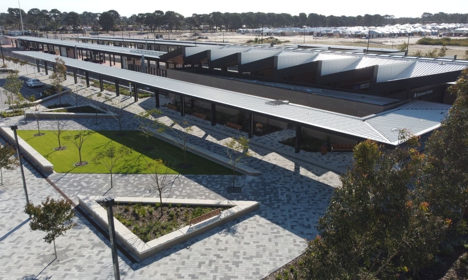 A modern train station with covered platforms, landscaped walkways, and a parking lot, with trees and buildings in the background.