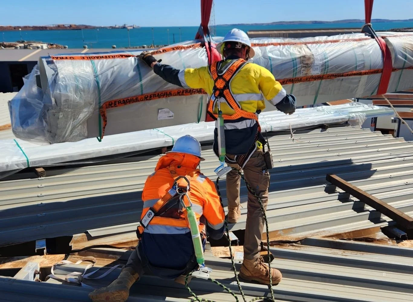 Two construction workers in safety gear installing a large wrapped component on a metal roof at a construction site near the water.