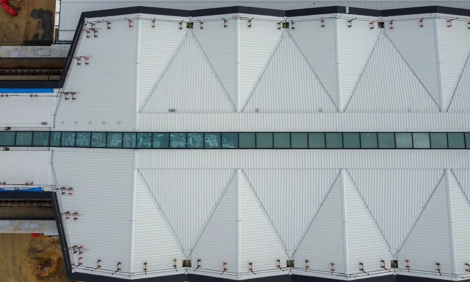 Top view of a building's roof with white panels, a horizontal row of windows, and construction materials and ground visible around the edges.