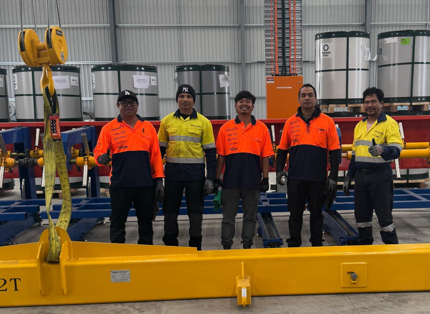 Five workers in safety gear standing inside an industrial warehouse. They are dressed in high-visibility shirts, some orange, some yellow, with safety gloves. Behind them are large metal tanks, and to the side is a yellow overhead crane hook.