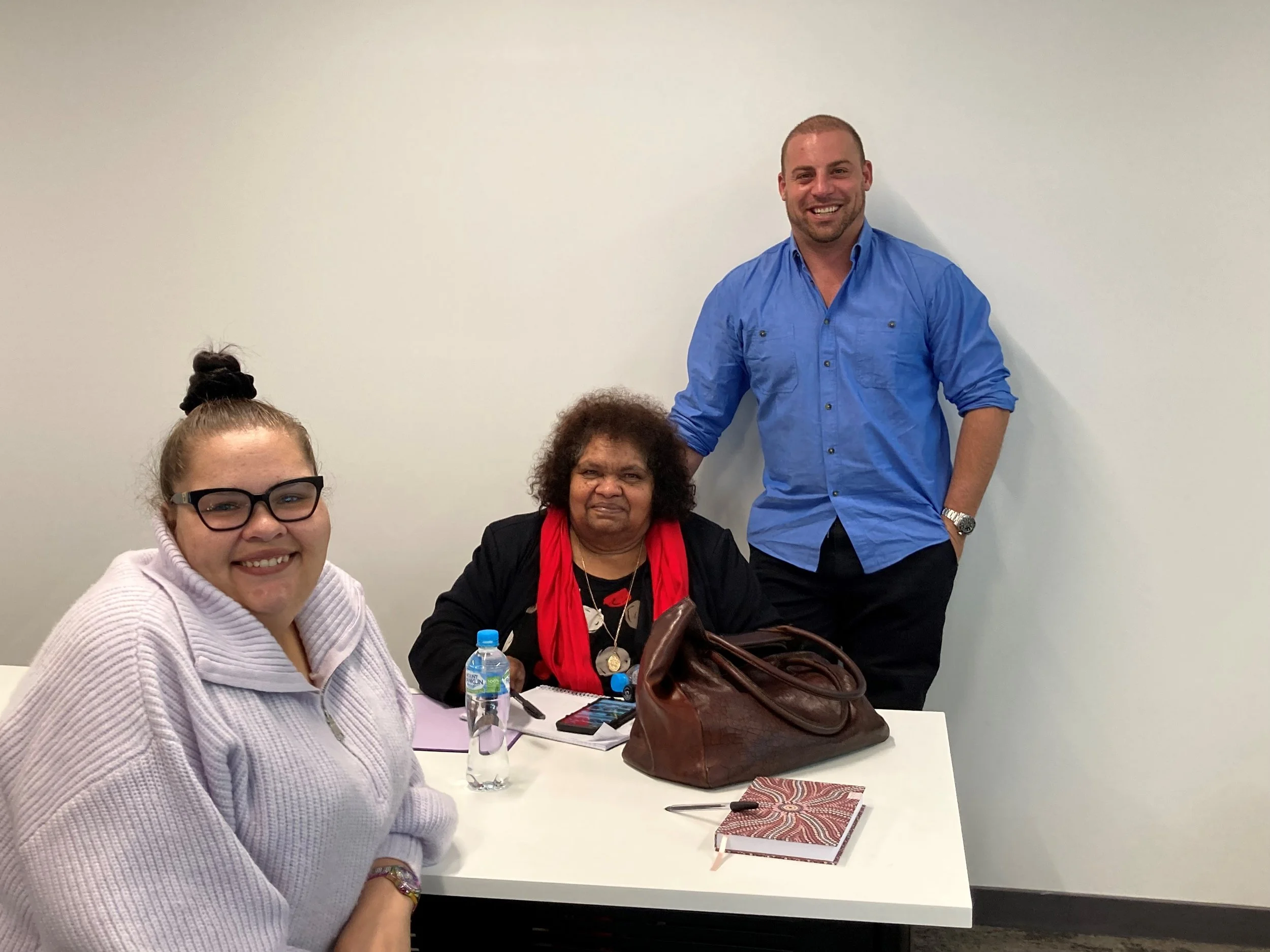 Four people are gathered around a white table in a room with white walls. One woman wearing glasses and a light-colored sweater is smiling at the camera. Next to her, a woman with dark curly hair, wearing a black jacket and red scarf, is seated with 