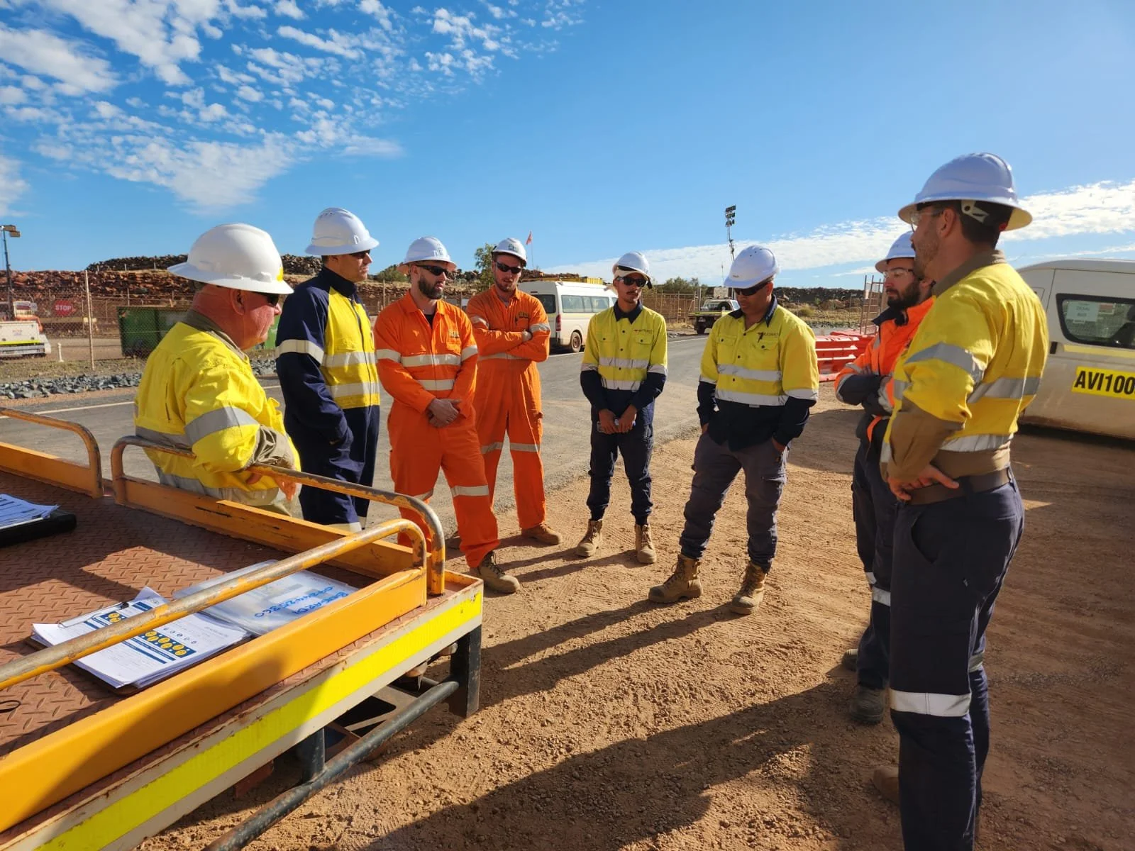 Construction workers and supervisors in safety gear having a discussion outdoors at a construction site under a blue sky with scattered clouds.