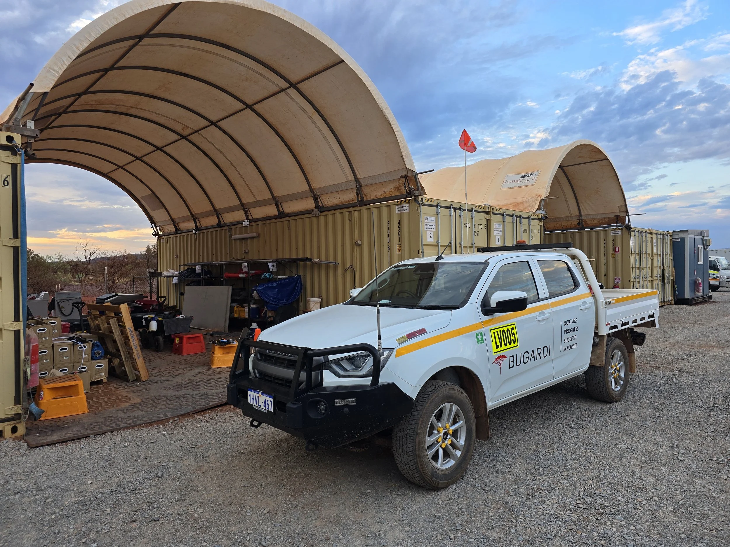 A construction or work site with a large arch-shaped fabric cover, several yellow storage containers, and a white utility vehicle with logos and text on it. The background shows a partly cloudy sky and some trees.