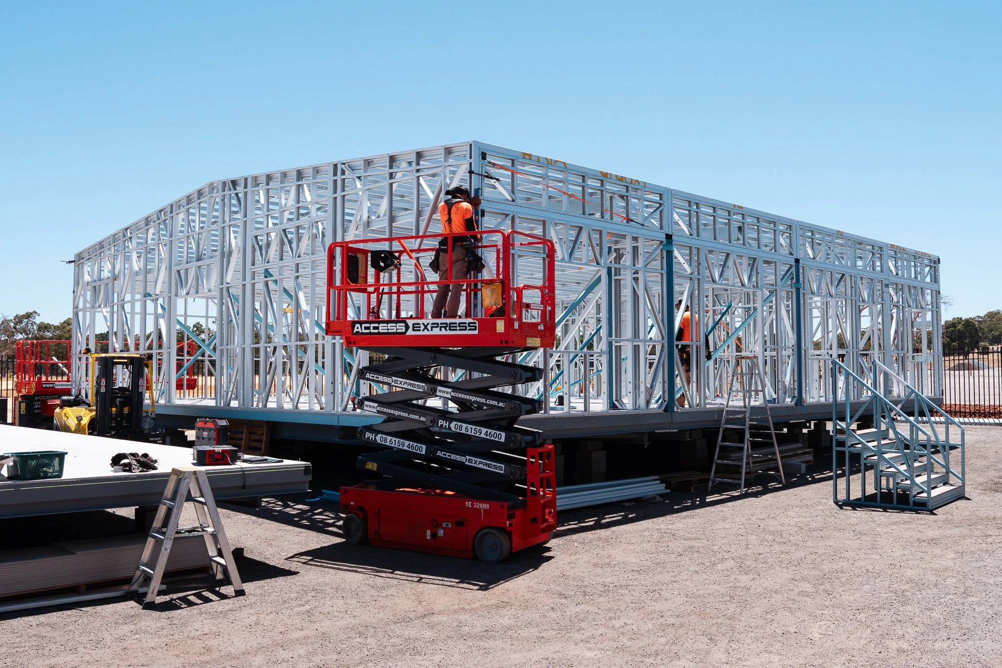 Construction workers assembling a metal framework on a raised platform outdoors, with various tools and equipment nearby under a clear blue sky.