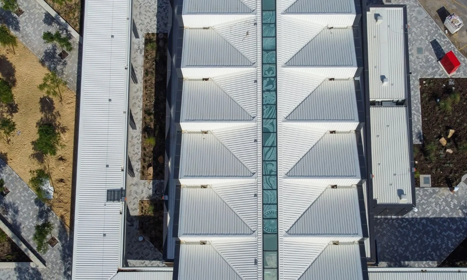 Aerial view of a large building with a white metal roof featuring a decorative glass walkway in the center, surrounded by paved walkways, trees, and landscaping.