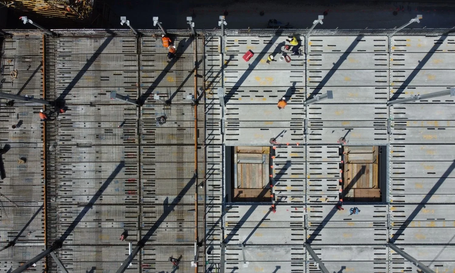 An aerial view of a construction site showing workers, scaffolding, and metal framework for a building. Two large square openings are visible in the structure.