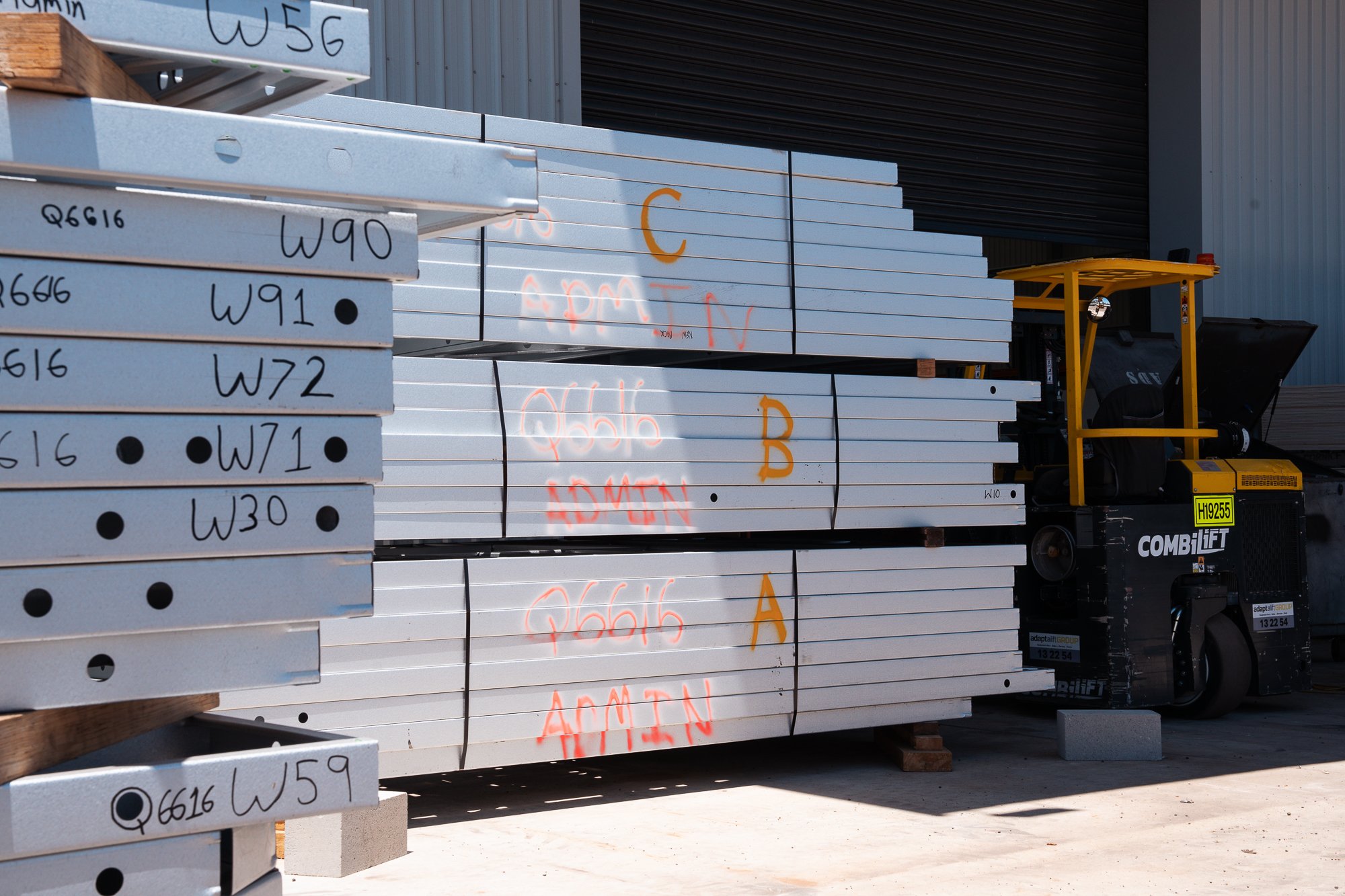 Stacked metal beams with handwritten labels, in front of a warehouse exterior, with a yellow lift machine on the right side.