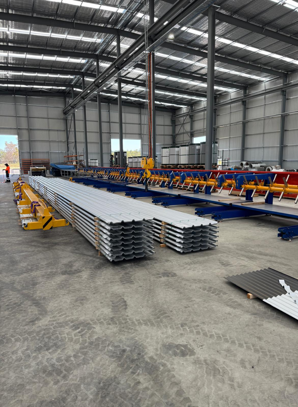 Interior of a metal fabrication warehouse with stacks of metal beams and panels, overhead cranes, and industrial equipment.