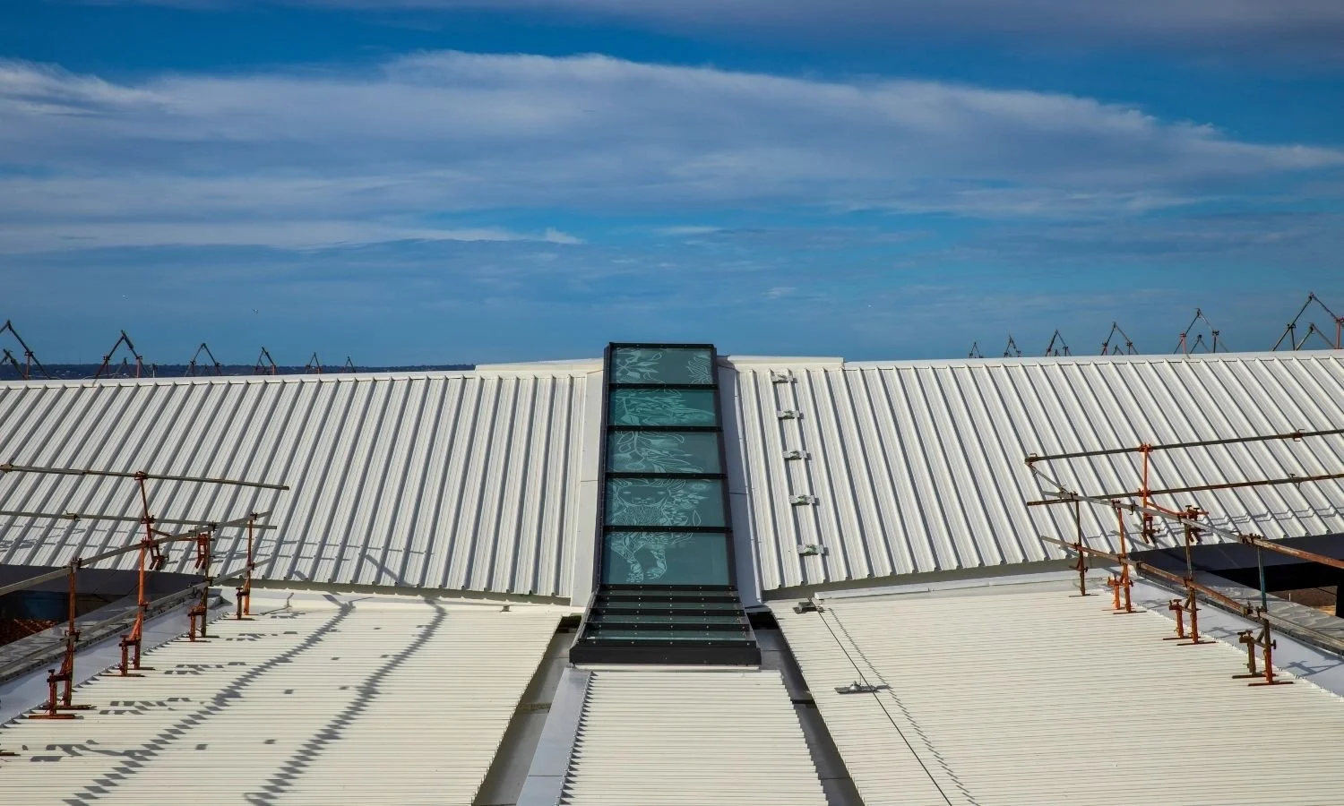 View of a white rooftop with a glass skylight in the center, under a partly cloudy sky, with cranes in the background.