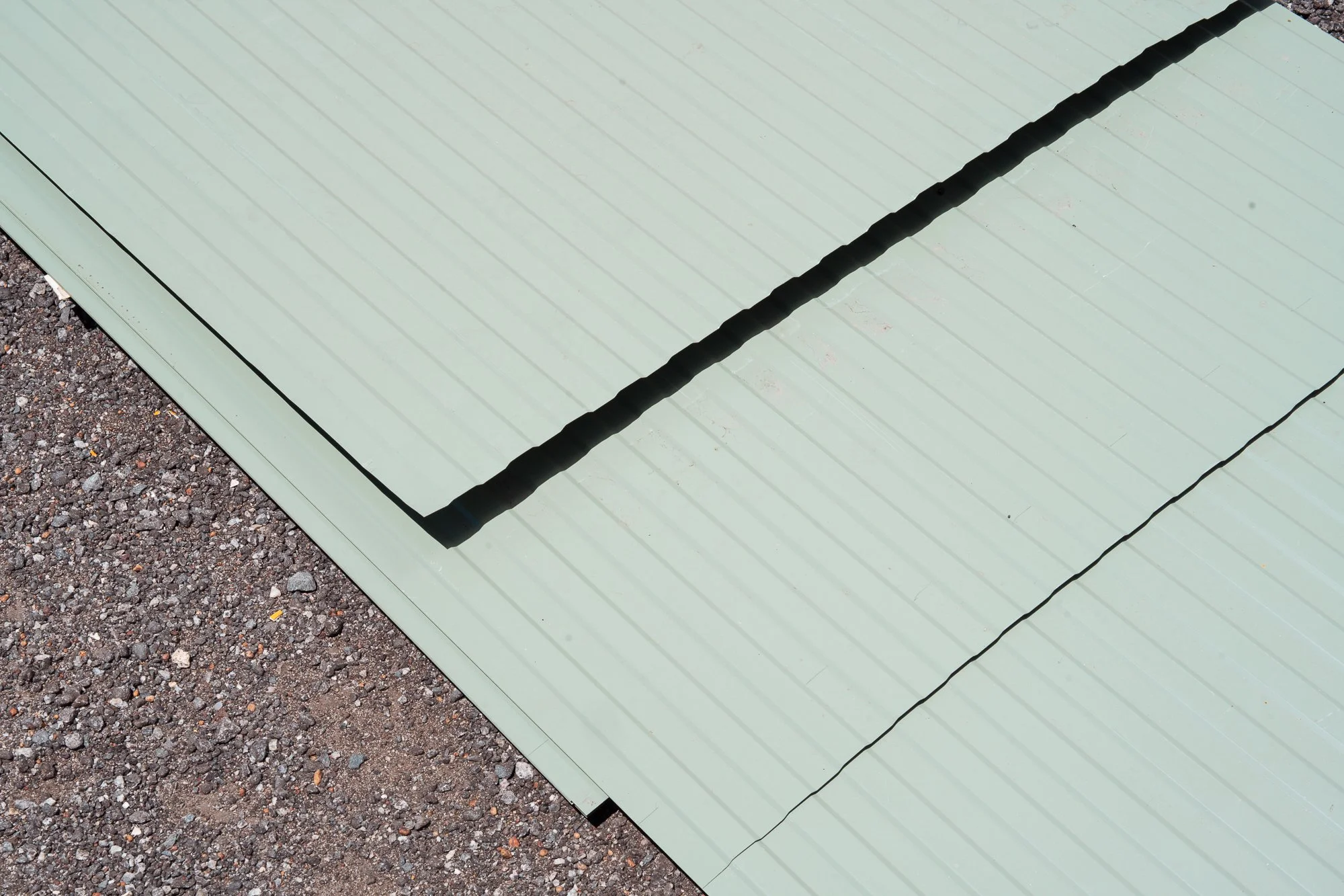 Close-up of light green metal roof panels with a black seam, surrounded by gravel ground.