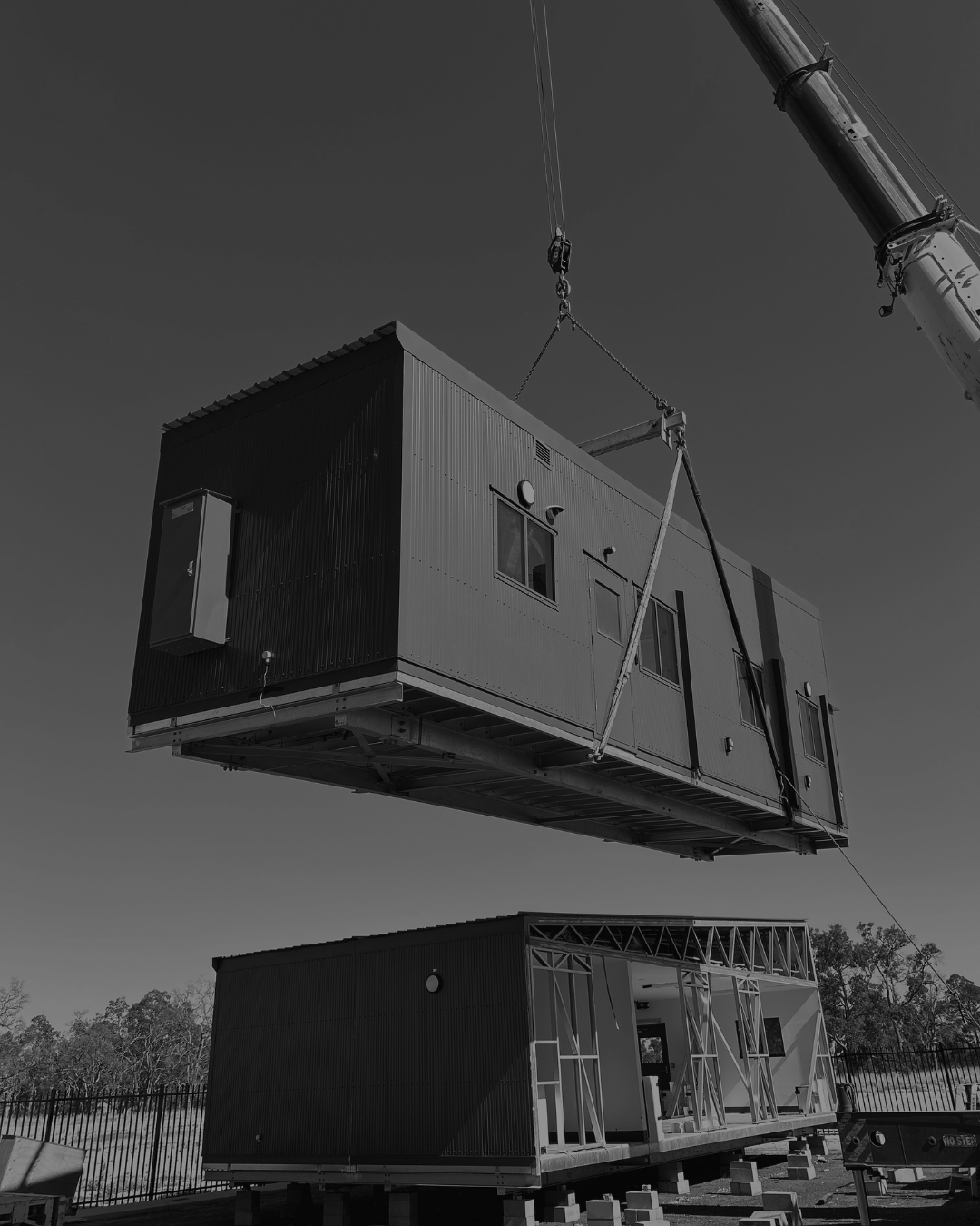 A crane lifting a prefabricated modular building above another similar building in an outdoor construction site.