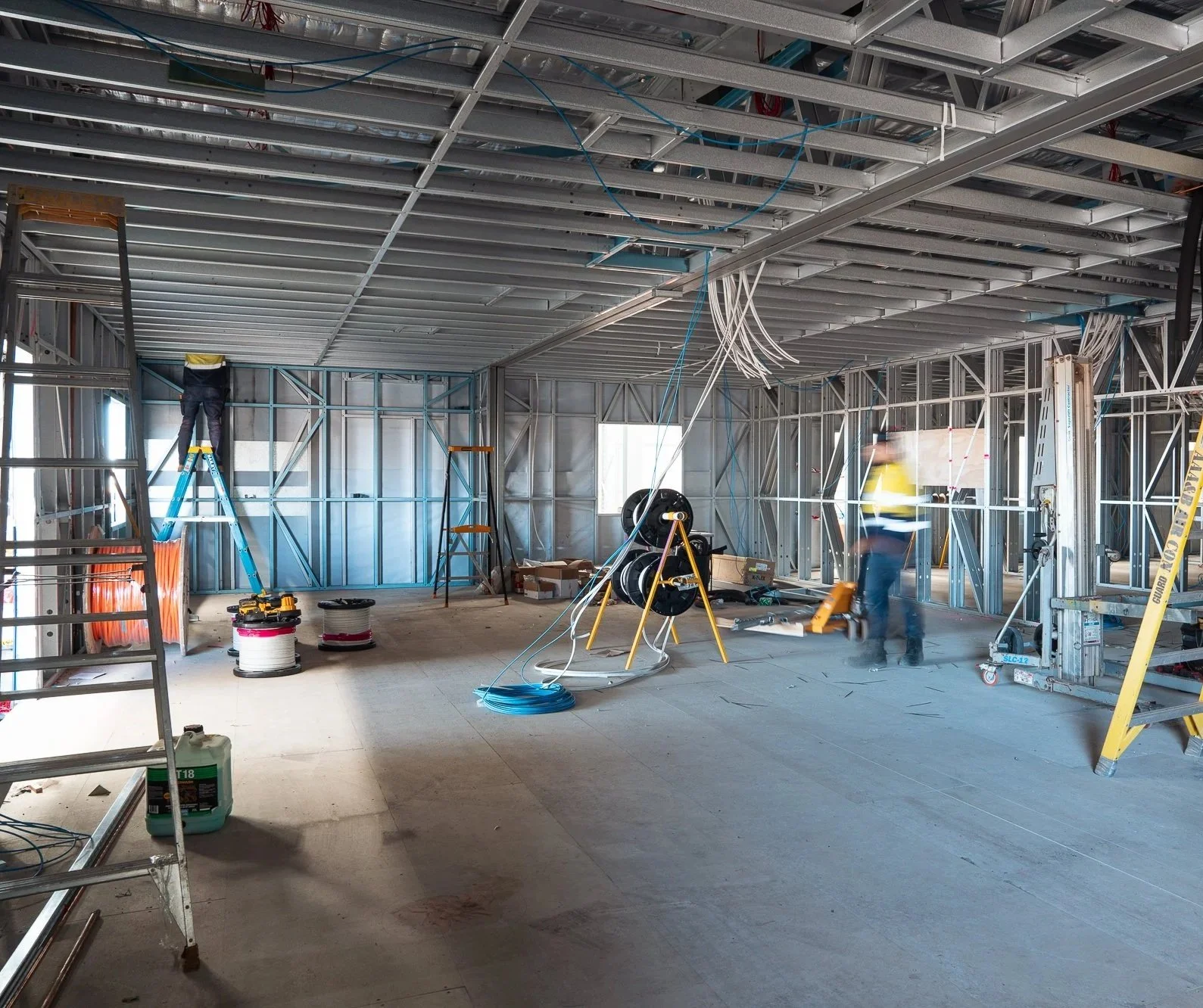 Interior of a building under construction with metal framing, construction workers, and various construction tools and materials, including ladders, wiring, and spool of cables.