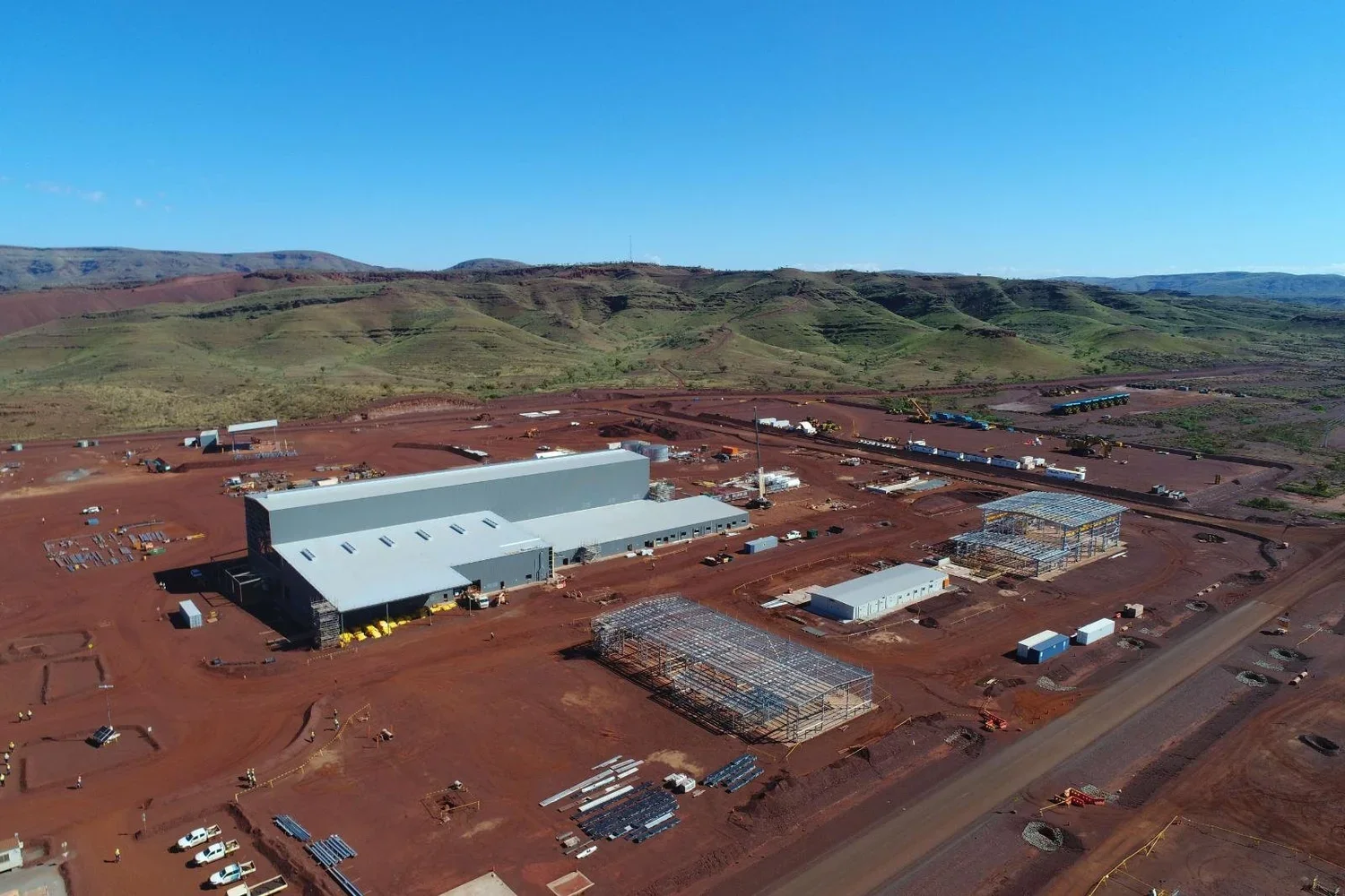 Aerial view of a construction site in a desert, with large buildings, scaffolding, construction vehicles, and equipment, surrounded by red soil and green hills in the background.