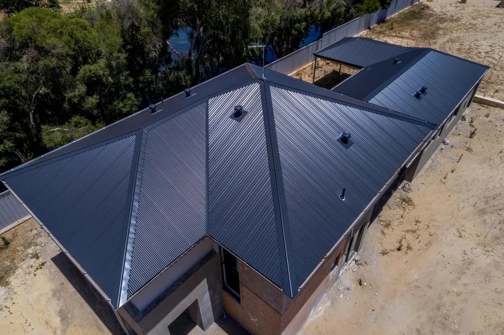 Aerial view of a modern house with a dark metal roof, situated near trees and an open yard.