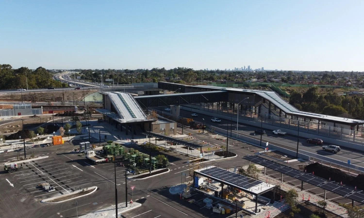 An aerial view of a modern transit station under construction with parking lots, roads, and a city skyline in the background.