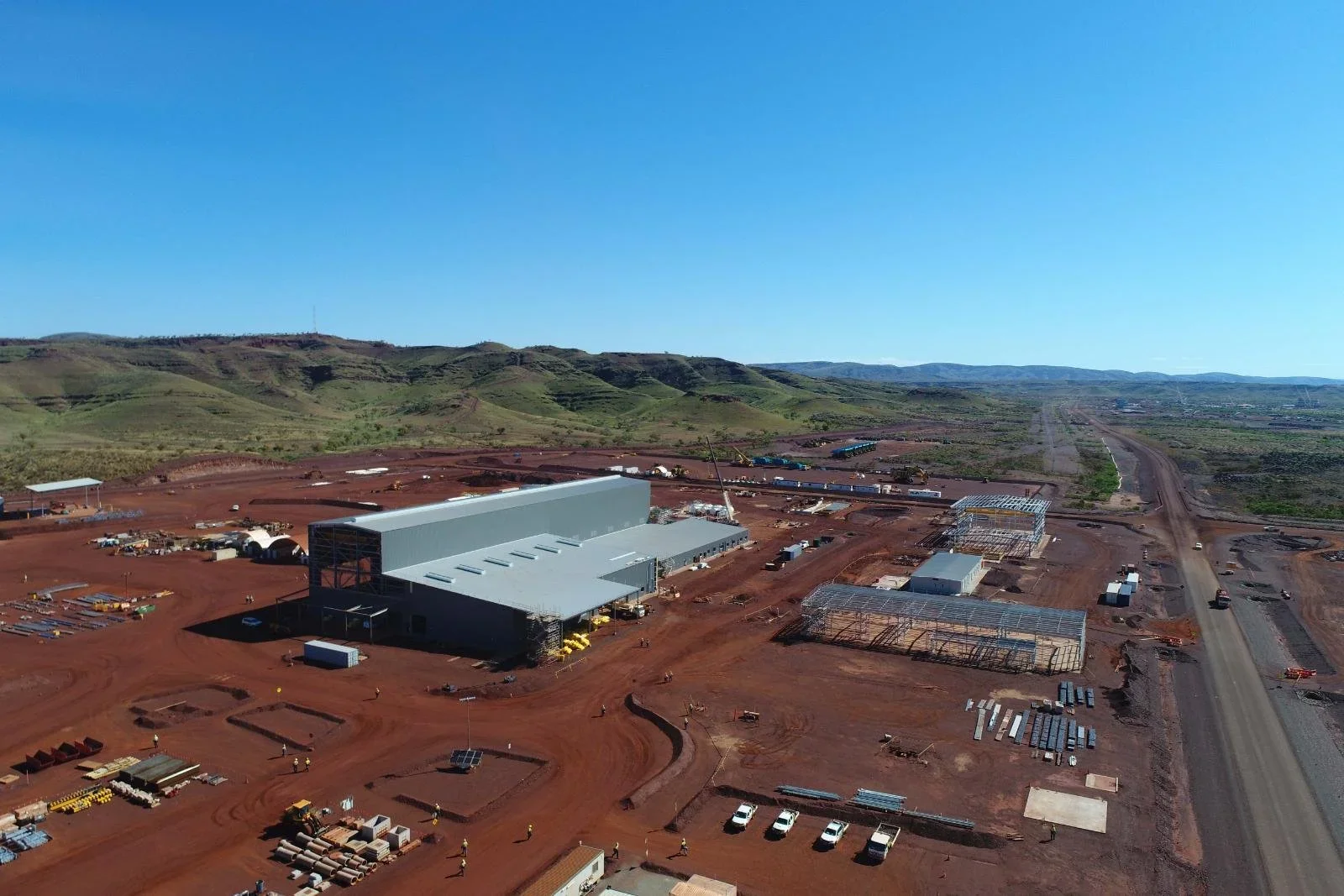 Construction site with a large gray building under construction, surrounded by dirt, equipment, and framed structures in a rural area with hills and open land