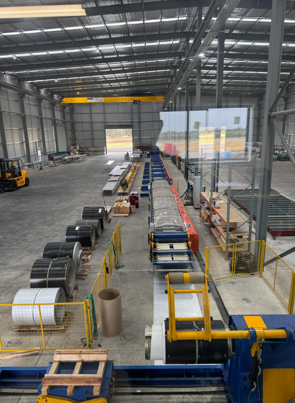 Interior of a large industrial warehouse with metal walls and ceiling. There are industrial machines, steel coils, workbenches, a forklift, and a yellow overhead crane. The space is organized with various equipment and materials for manufacturing or 