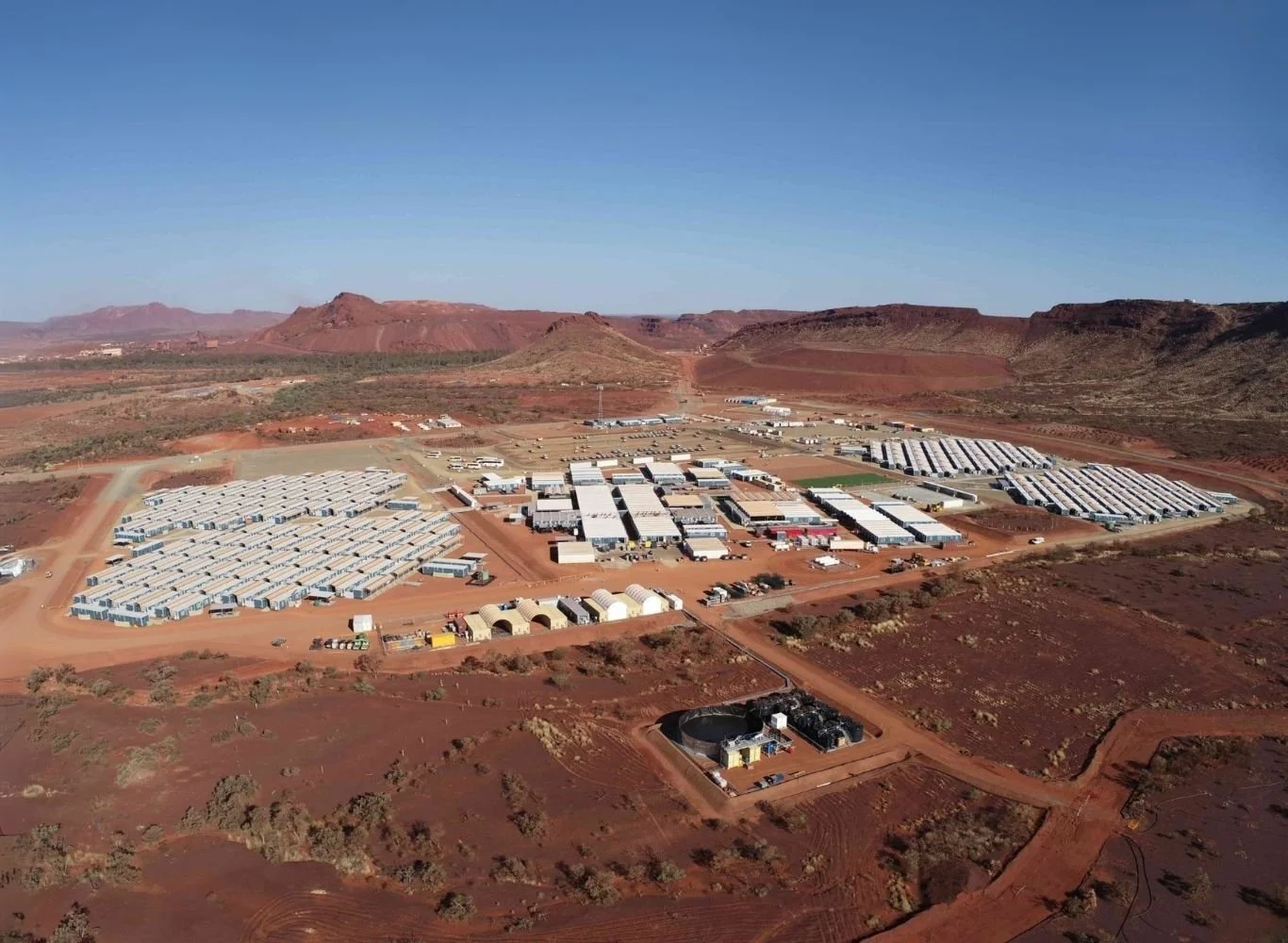 Aerial view of a large facility in a desert landscape with mountains in the background, featuring numerous white temporary buildings, vehicles, and dirt roads.