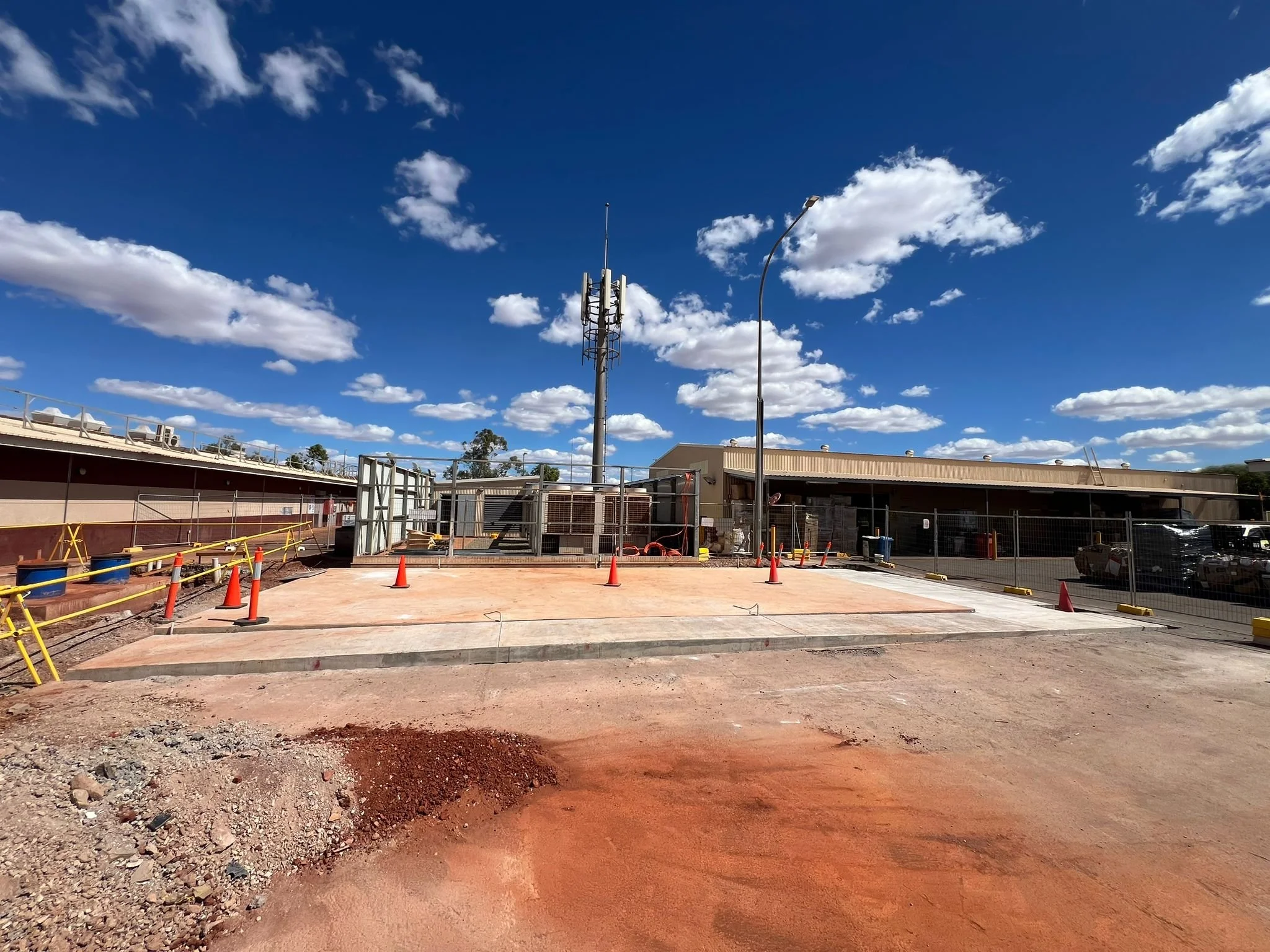 Construction site with orange gravel and orange cones, fenced area, with a building in the background, tall communication tower, and blue sky with clouds.