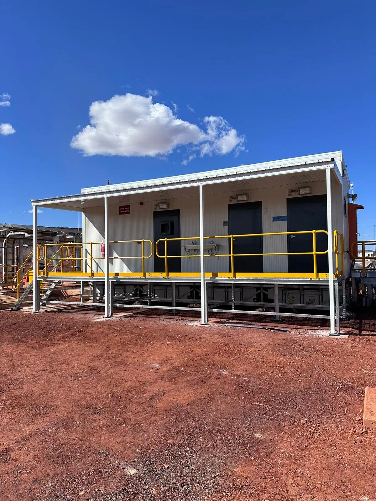 A small white metal building on a raised platform with a yellow safety railing, set against a bright blue sky with scattered white clouds.